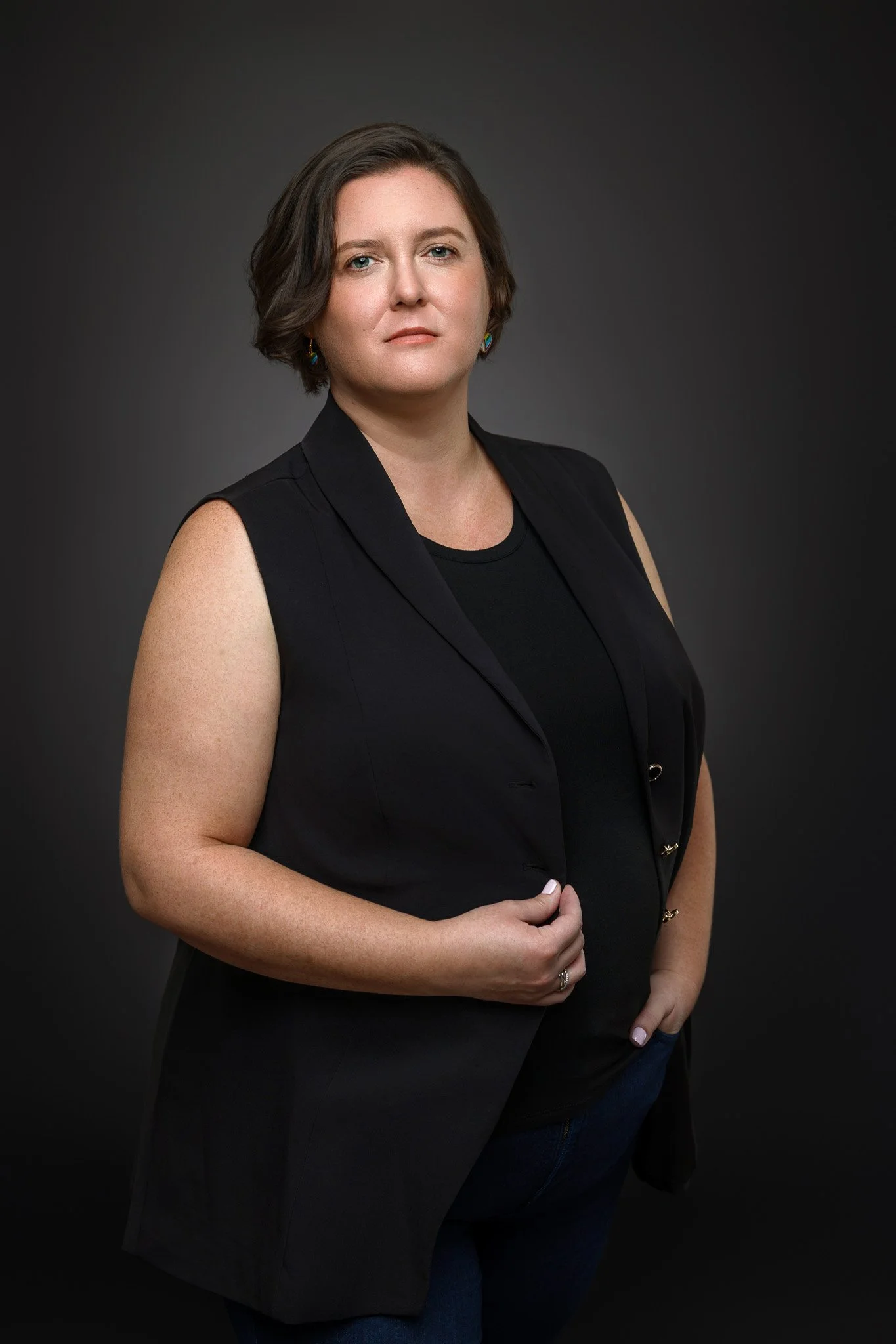 Portrait of a woman with short brown hair wearing a black sleeveless blazer and a black top, standing against a dark gray background.