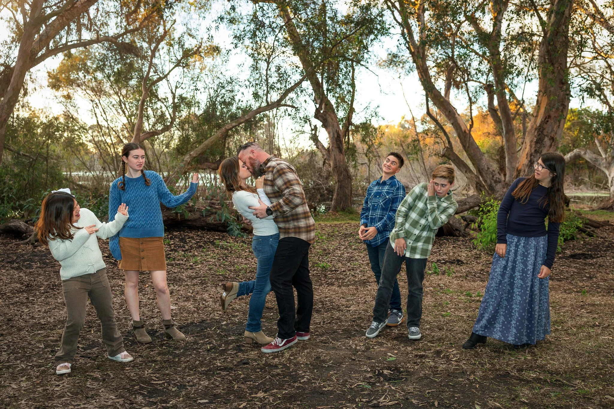 Family of seven outdoors in a wooded area during sunset, with a man and woman kissing in the center and children around them making playful gestures.