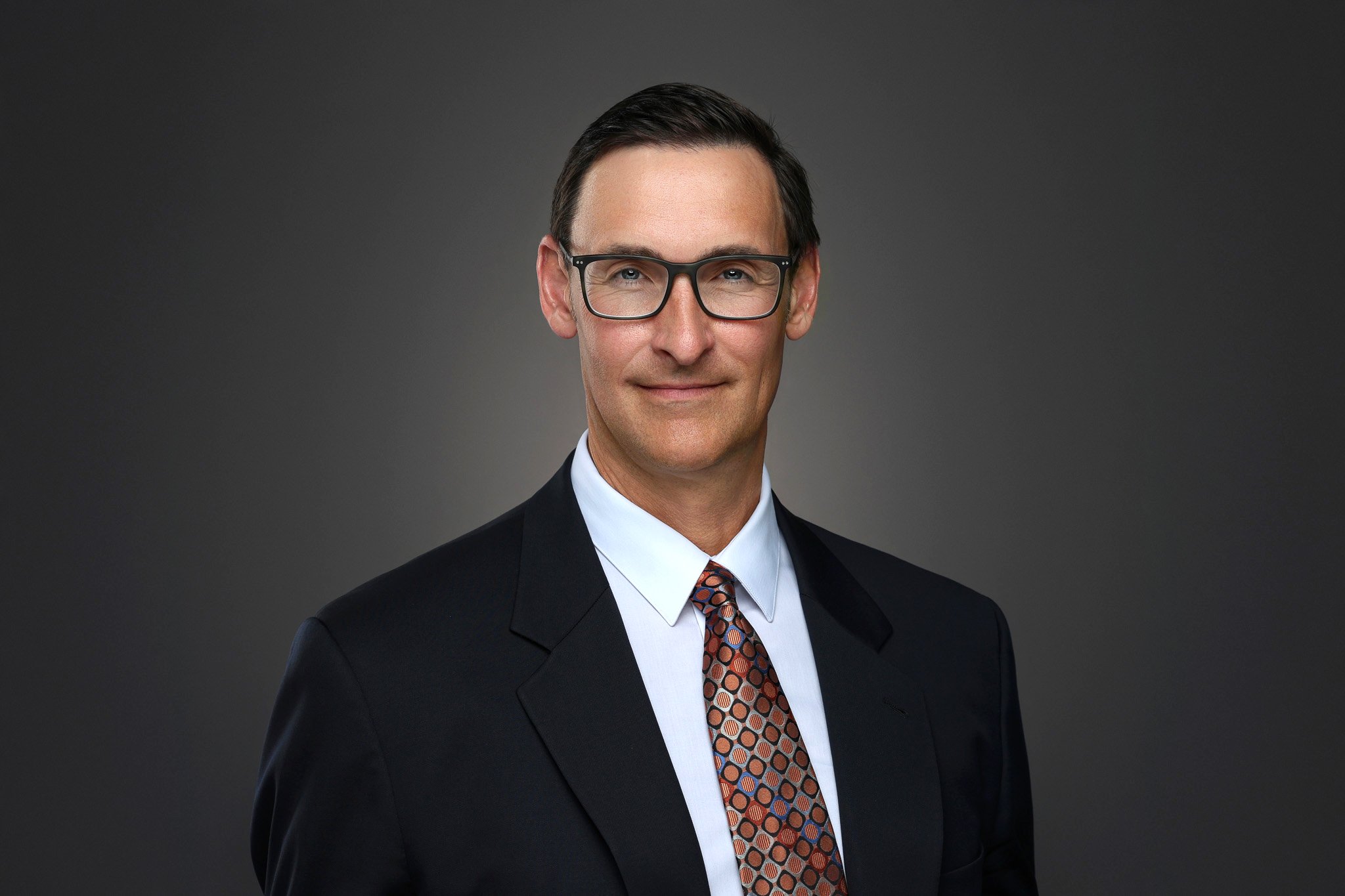 A middle-aged man in a black suit, white shirt, patterned tie, wearing black glasses, smiling slightly, against a gray background.
