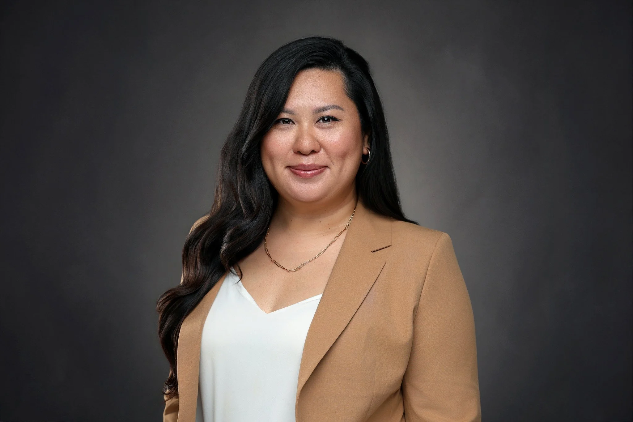 A professional woman with long dark hair, wearing a tan blazer over a white top, smiling against a dark gradient background.