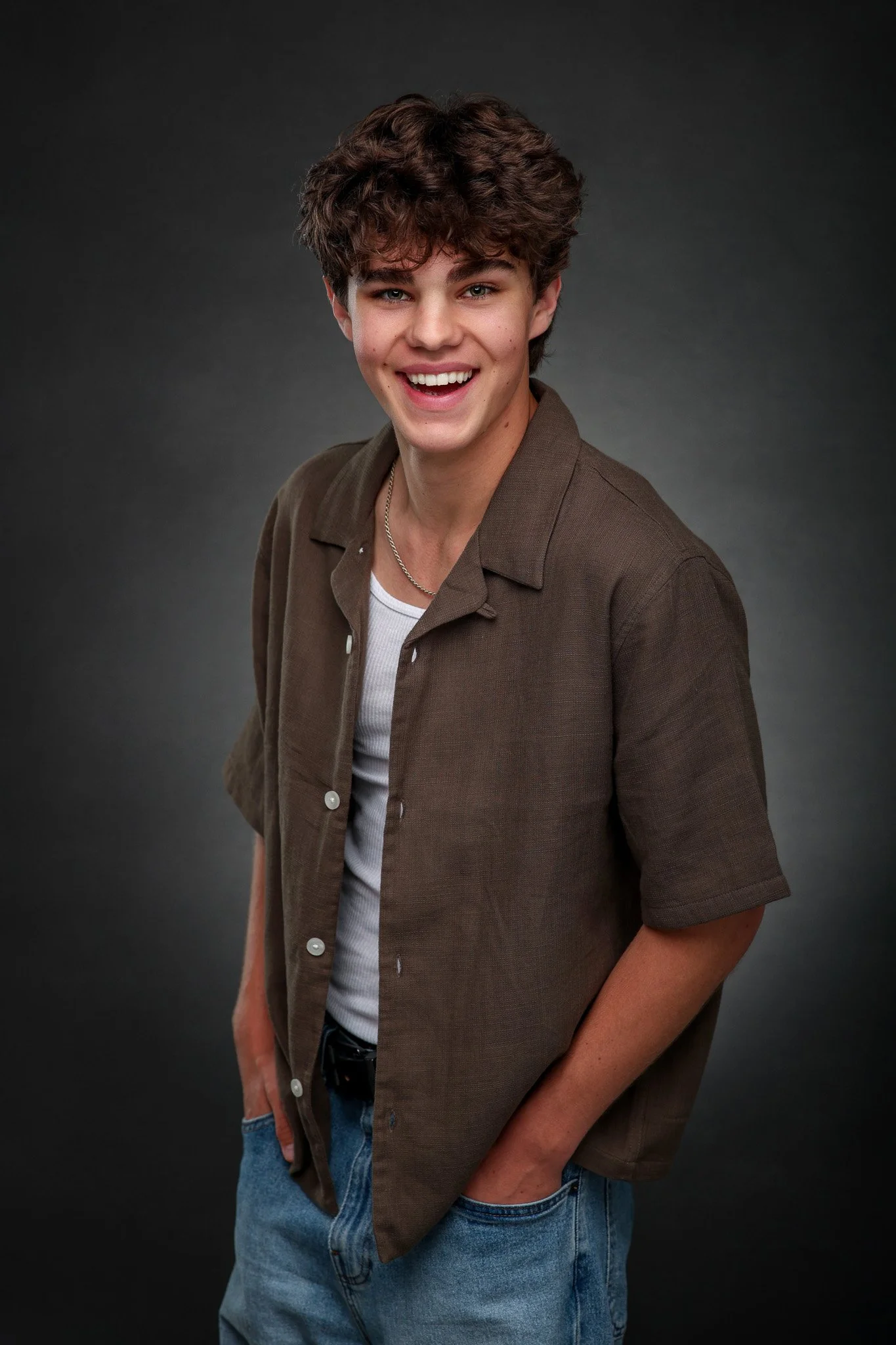 A young man with curly brown hair smiling, wearing a brown button-up shirt over a white t-shirt and blue jeans, standing against a dark gray background.