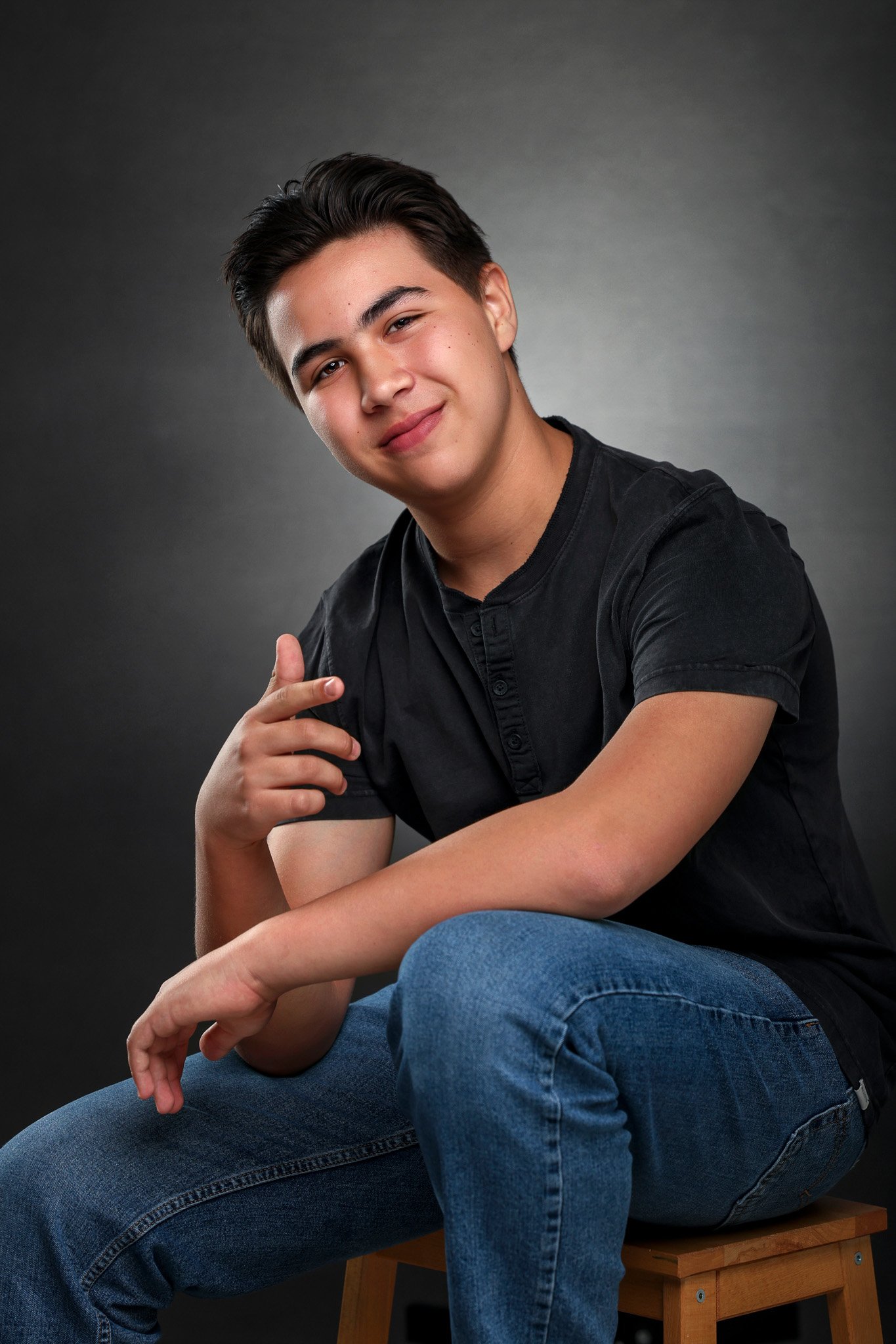 A young man with short dark hair, smiling, sitting on a wooden stool against a gray background, wearing a black t-shirt and blue jeans, pointing with his right hand.