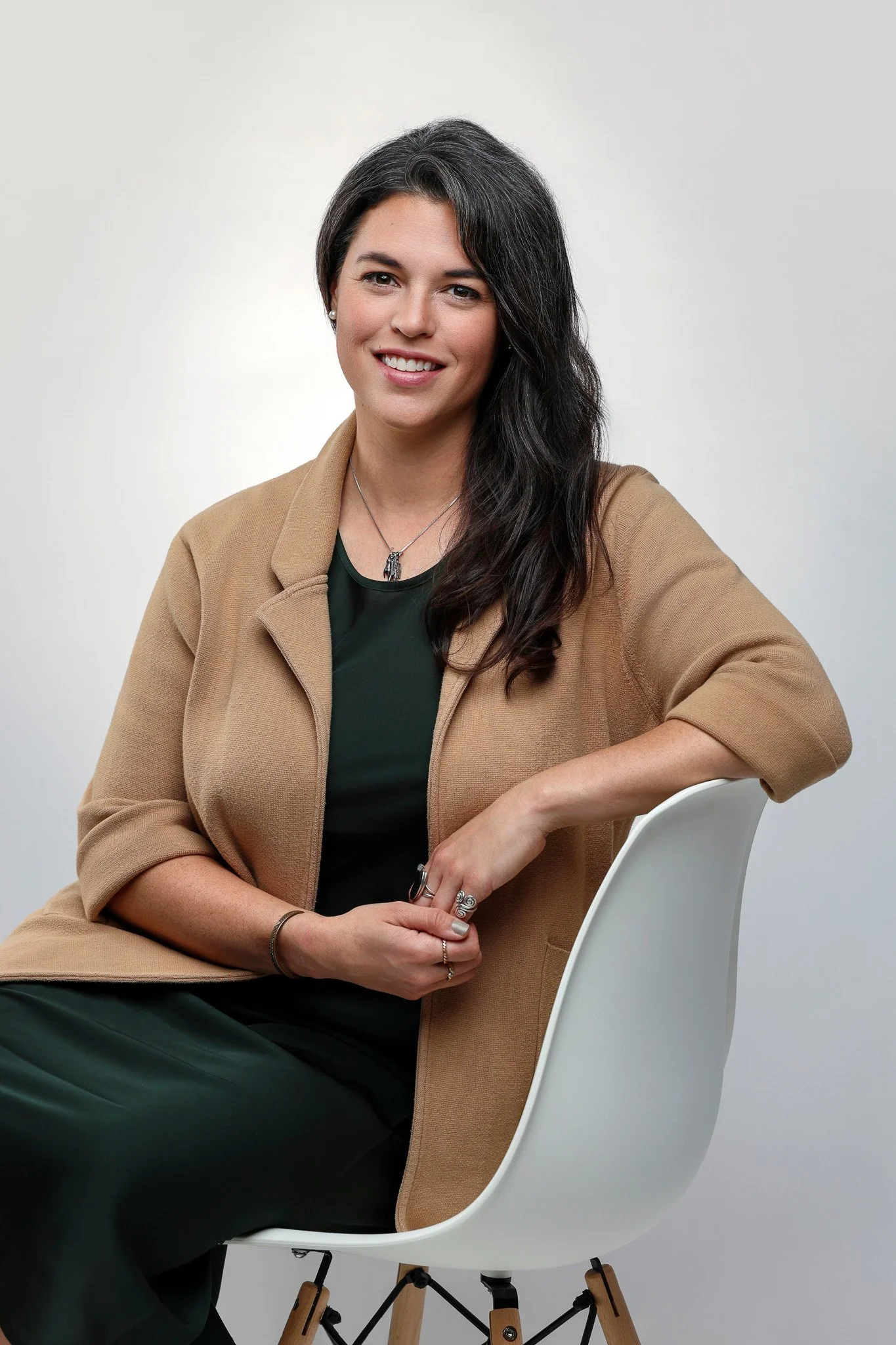 A woman with long dark hair, wearing a tan coat over a dark top, sitting on a white chair with wooden and metal legs, smiling at the camera against a plain light background.