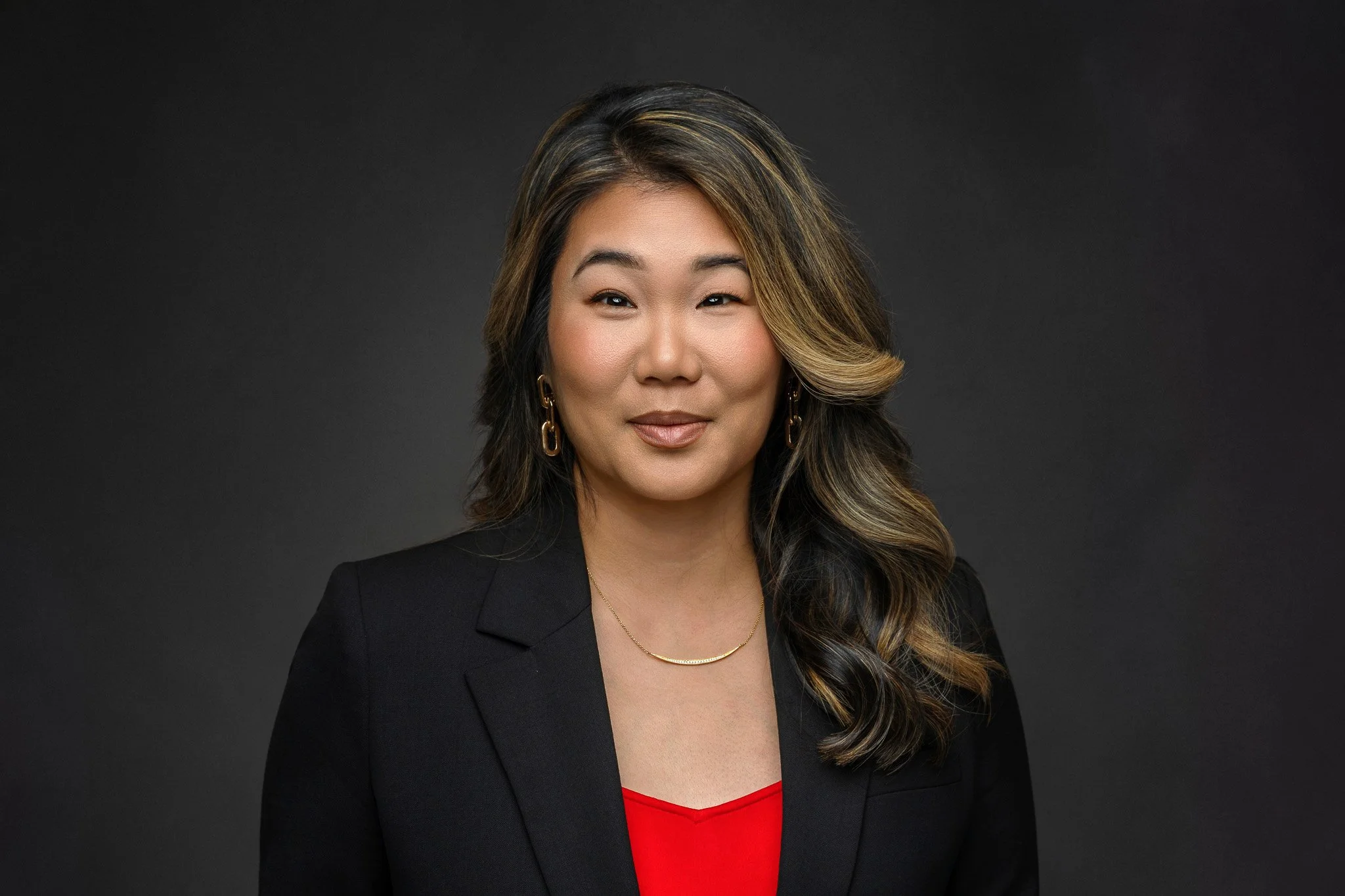 Professional woman with long, wavy hair, middle part, wearing a black blazer over a red top and gold jewelry, posing against a dark gray background.