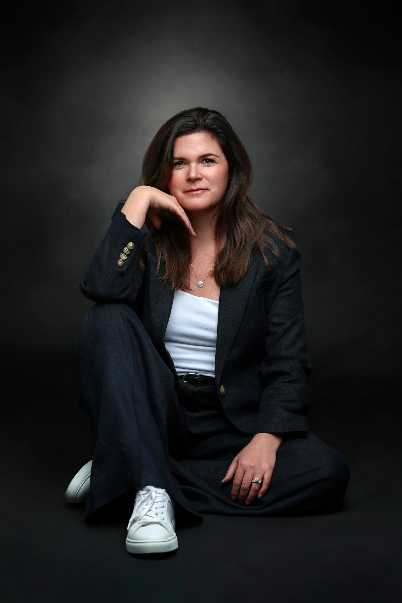 A woman with dark brown hair, wearing a black blazer and black pants, sitting on a black background, with one knee up and her hand resting on her cheek. She is wearing white sneakers, a silver necklace, and a ring on her right hand.