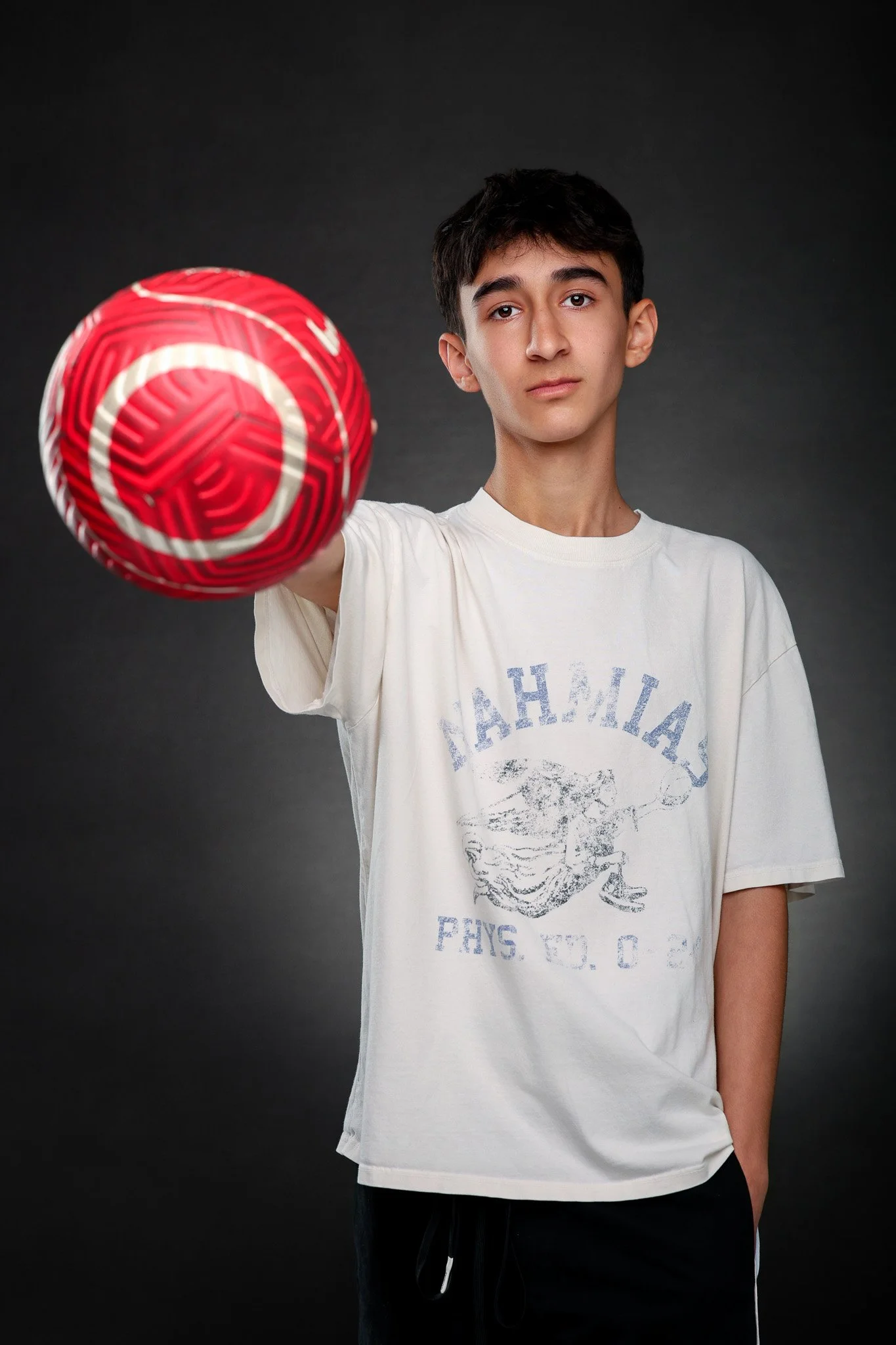 A young man with short dark hair is holding a red and white patterned volleyball in front of a dark background, looking directly at the camera.