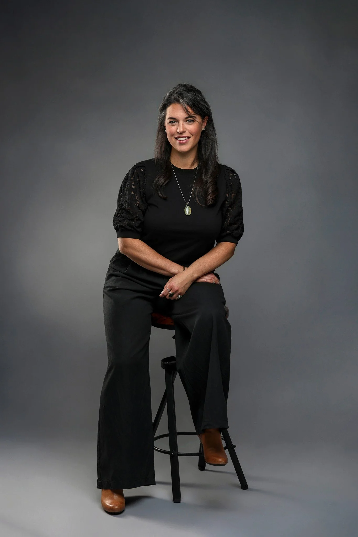 A woman sitting on a stool with a dark gray background. She has dark hair, is smiling, and is dressed in black with brown shoes, wearing a necklace.