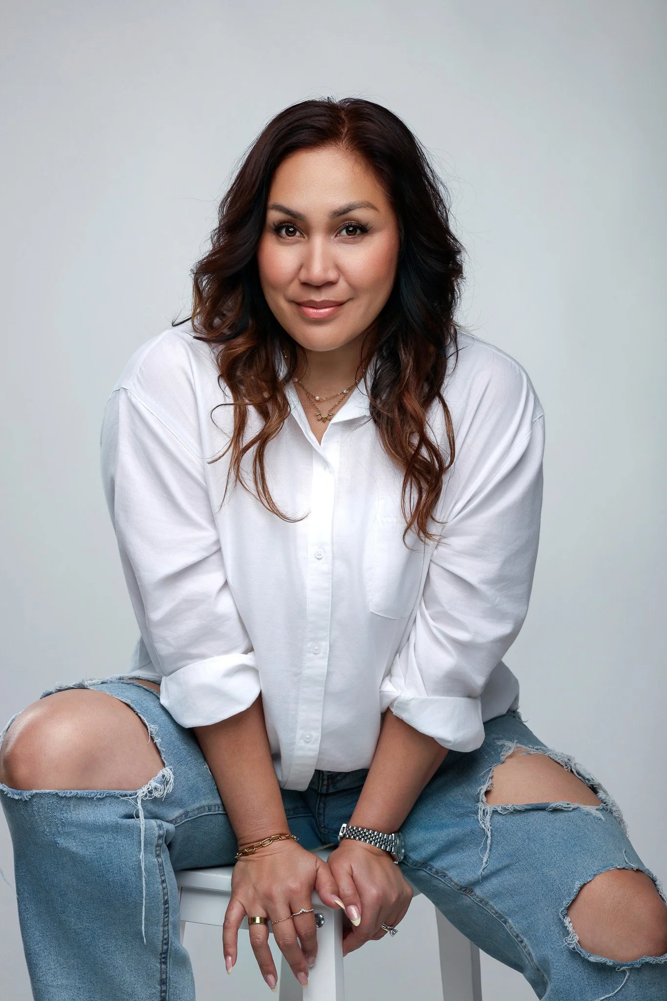 A woman with long wavy brown hair, wearing a white button-up shirt with rolled sleeves, ripped jeans, and jewelry, seated on a stool against a plain background.