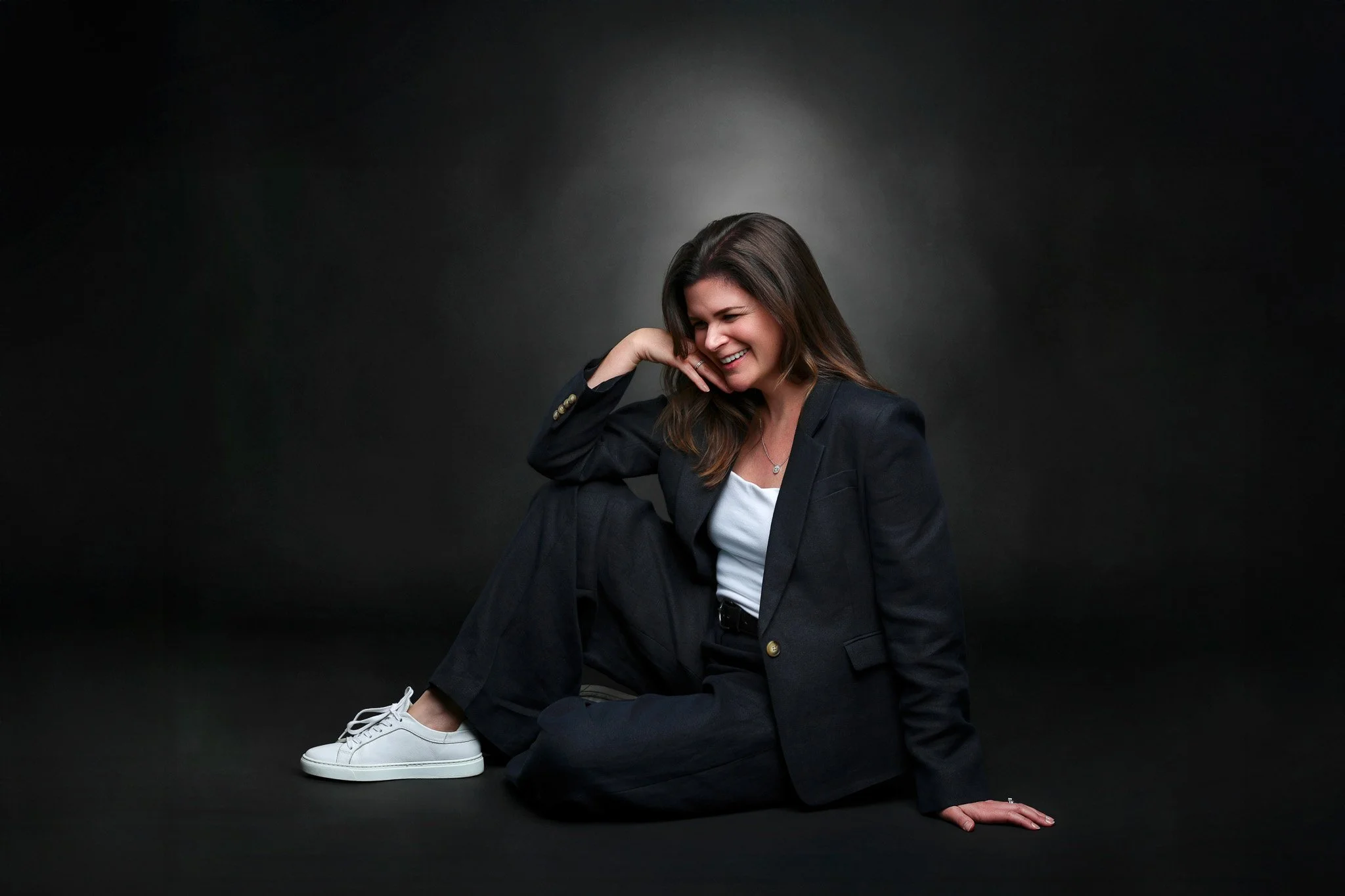 A woman with long brown hair sitting on the floor against a dark background, wearing a black suit and white sneakers, smiling and looking down.