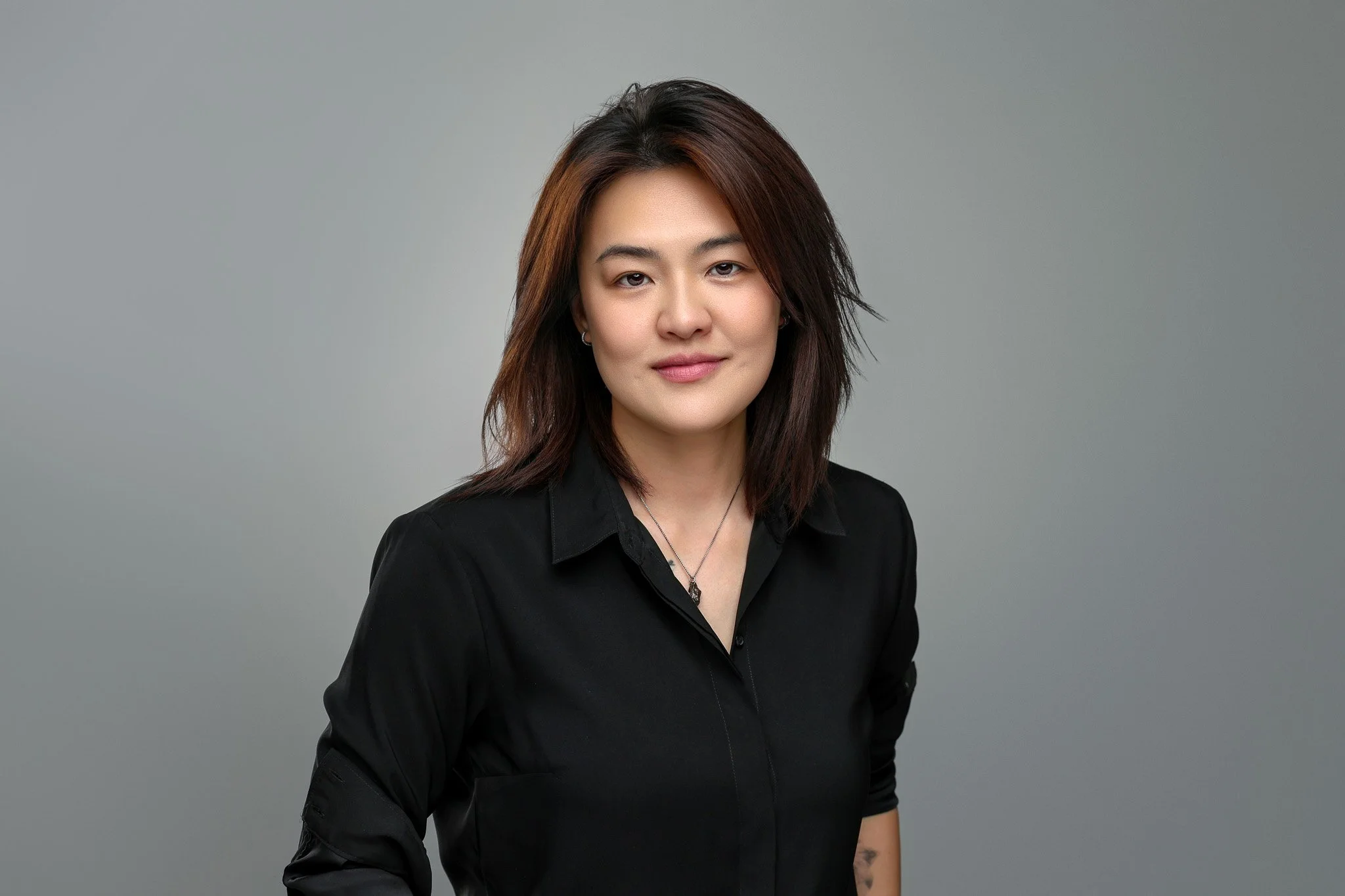 A woman with shoulder-length dark brown hair and light makeup, wearing a black shirt and necklace, standing against a plain gray background.