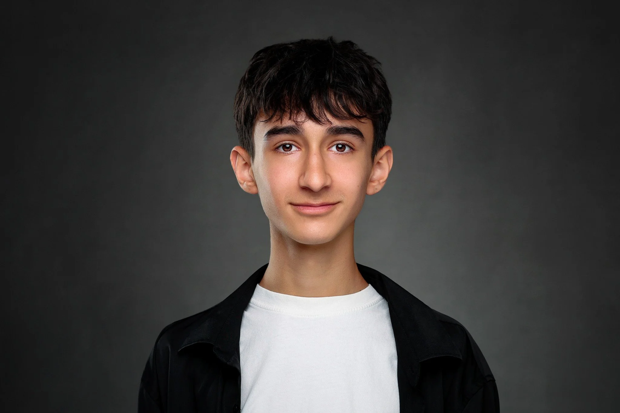 Young person with short dark hair, wearing a white shirt and black jacket, smiling lightly against a dark background.
