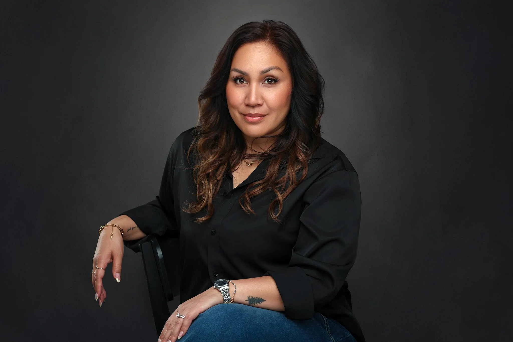 Portrait of a woman with long brown hair wearing a black shirt and blue jeans, sitting against a dark background