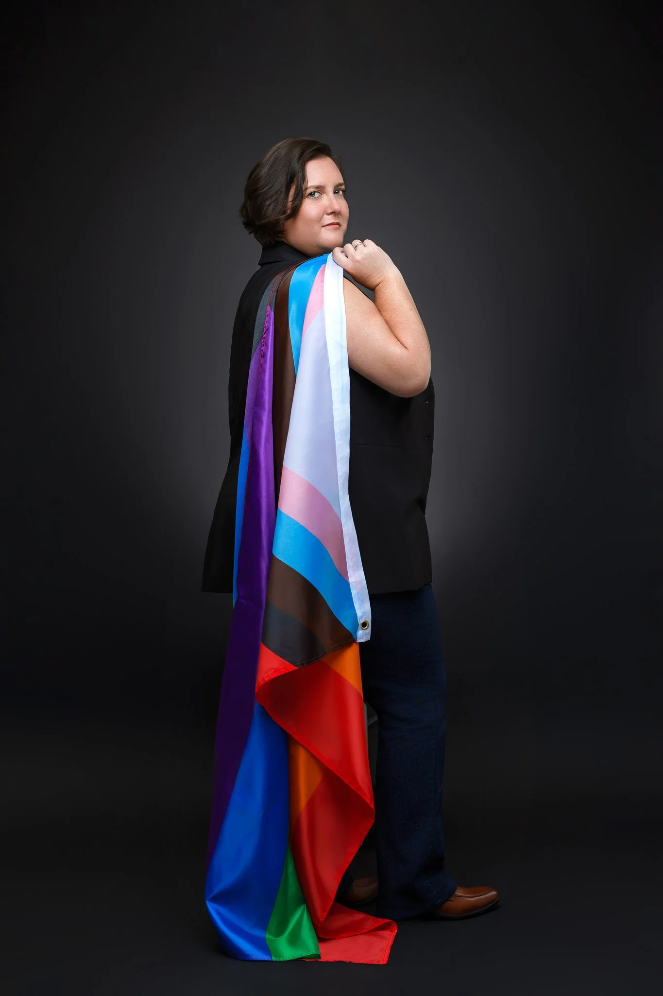 A person standing against a dark background, holding a rainbow pride flag over their shoulder and wearing a black blazer and jeans.