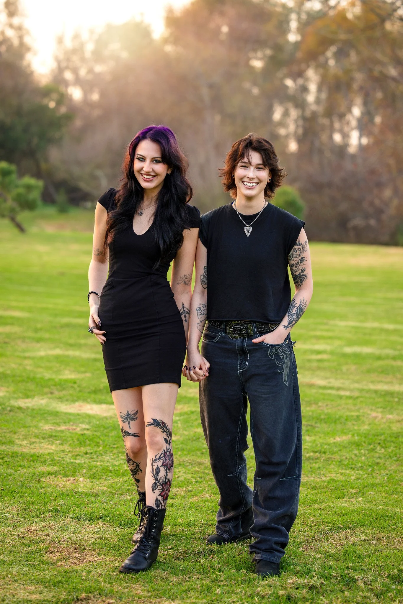 Two young women with tattoos and dark hair smiling and holding hands outdoors in a park during sunset.