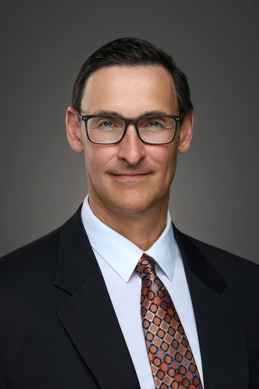 Professional portrait of a middle-aged man with dark hair, wearing glasses, a white shirt, a patterned tie, and a black suit against a grey background.