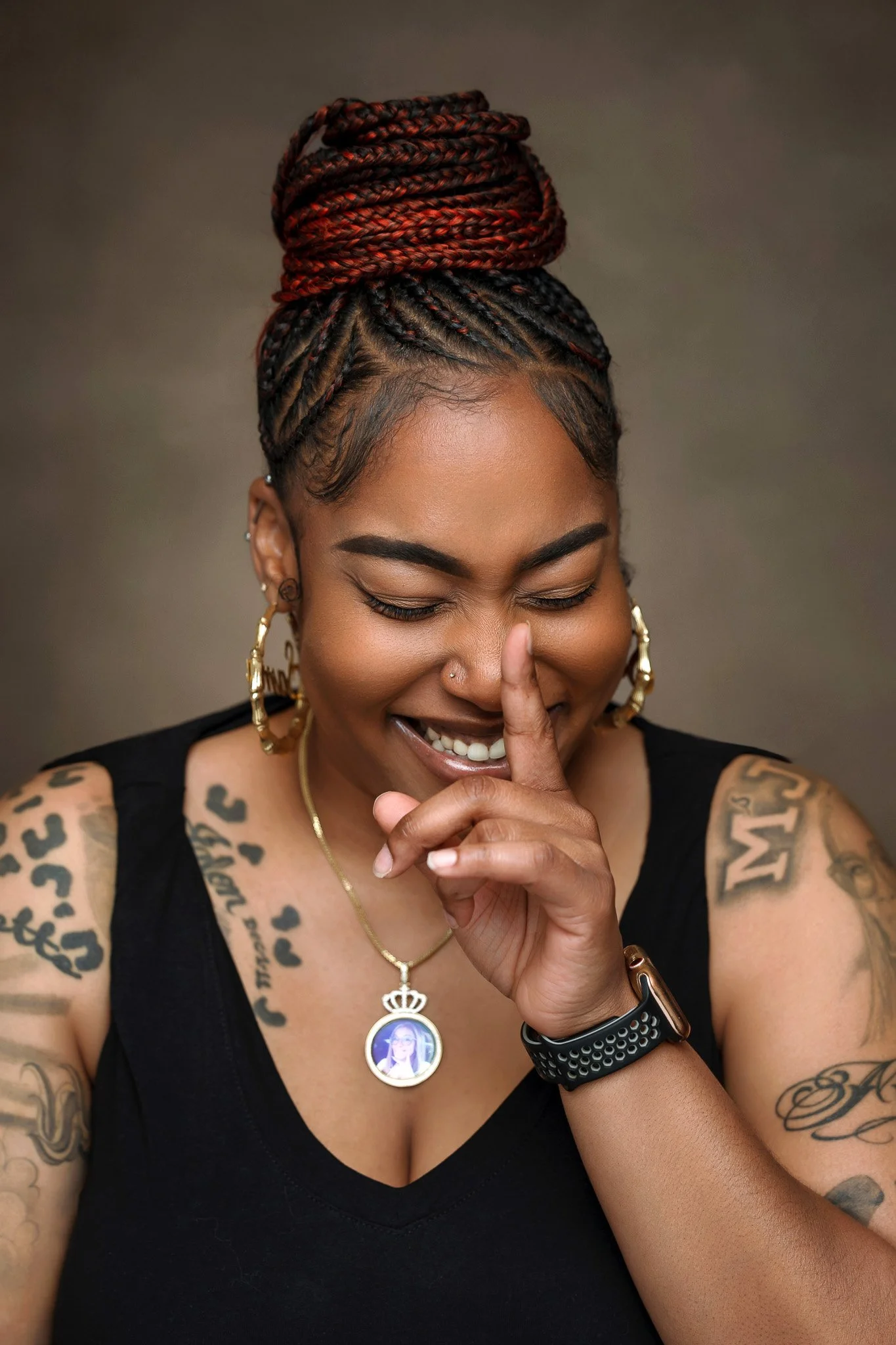 A woman with dark skin and tattoos on her arms, smiling with her eyes closed and making a shushing gesture with her finger on her lips, wearing a black sleeveless top, gold earrings, and a necklace with a pendant showing a woman's face.