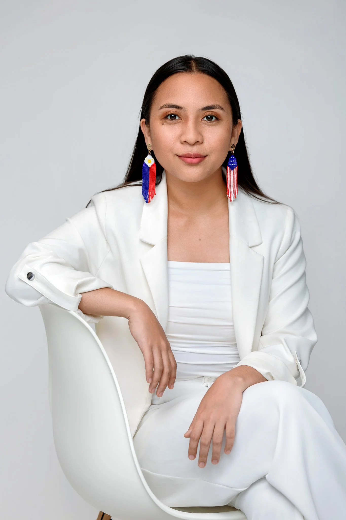 A woman with long dark hair sitting in a white chair against a plain light grey background. She is wearing a white outfit and colorful beaded earrings with red, white, blue, and yellow beads. She has a calm expression and is looking directly at the camera.