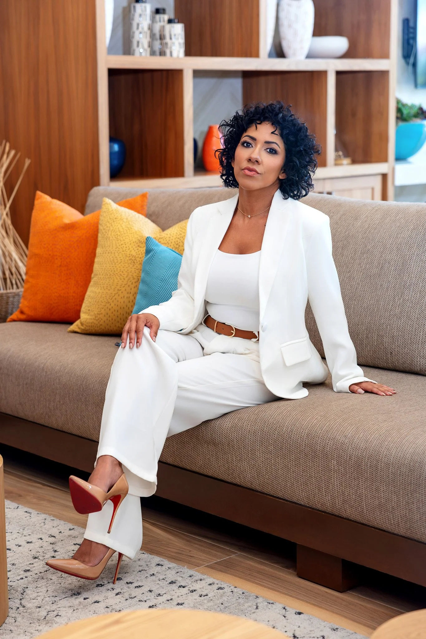 A woman with curly black hair and light brown skin tone sitting on a beige sofa in a modern living room, wearing a white suit with a brown belt, and beige high heels with red soles. She is looking at the camera with a confident expression, with color