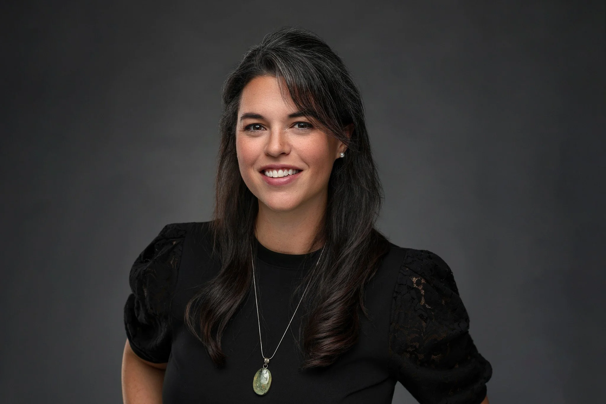 A woman with long, dark hair smiling, wearing a black top with lace on the shoulders and a pendant necklace, against a gray background.
