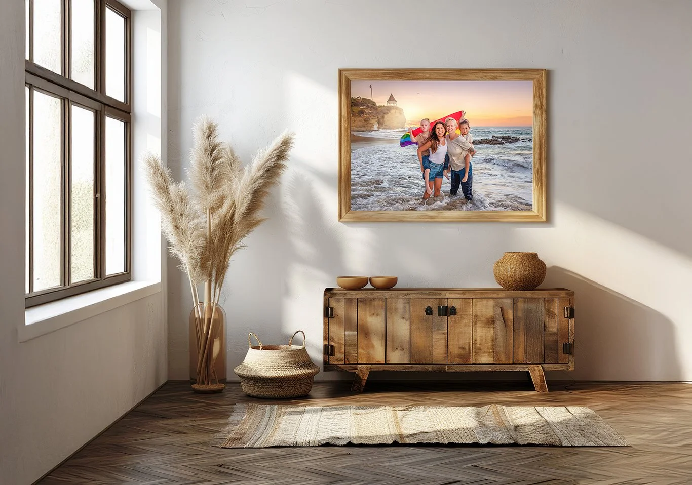 A cozy living room corner with a large framed beach photo on the wall, a wooden sideboard decorated with two bowls and a vase, a tall pampas grass arrangement, a woven basket, and a small rug on wooden flooring near a window letting in natural light.
