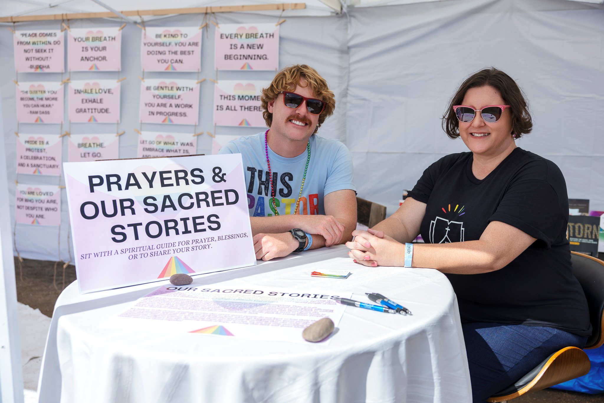 Two women sitting at a table with a sign that reads "Prayers & Our Sacred Stories" inside a white tent, smiling and holding hands, with colorful notes and posters in the background.