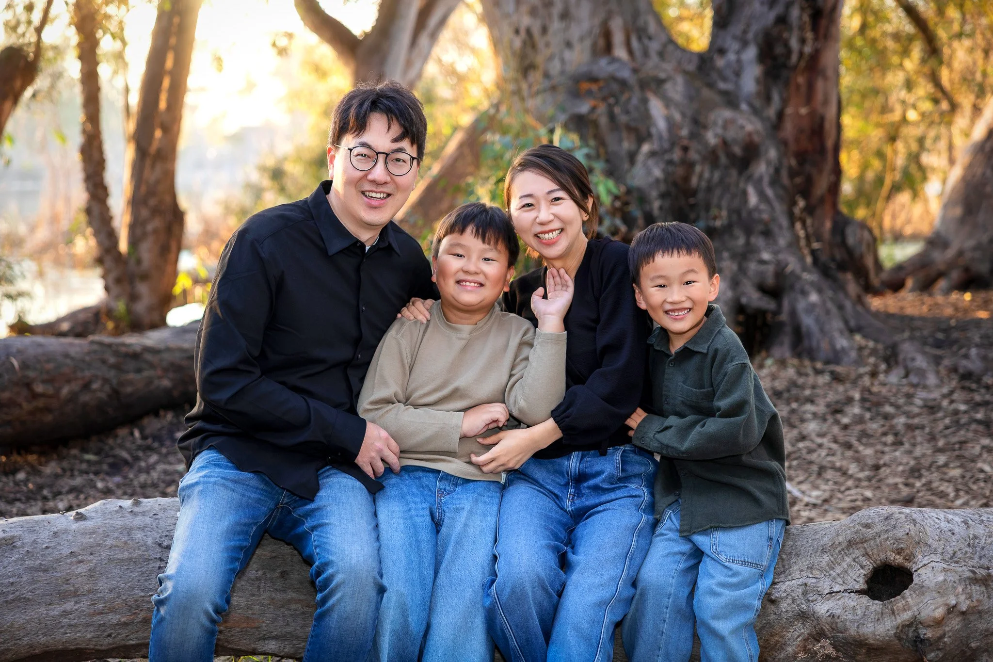 A happy Asian family of four sitting on a log outdoors in a park with trees and fallen leaves, smiling at the camera.