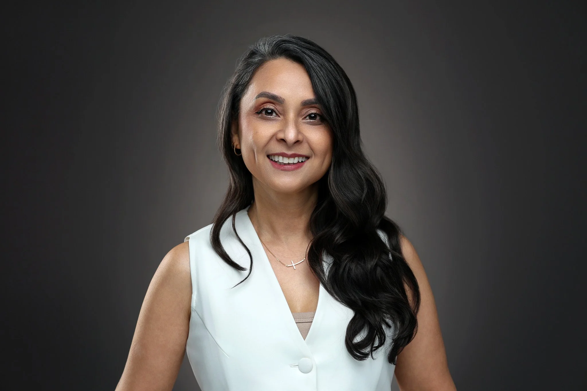 Portrait of a smiling woman with long dark hair, wearing a white sleeveless top and a gold cross necklace, against a dark gray background.
