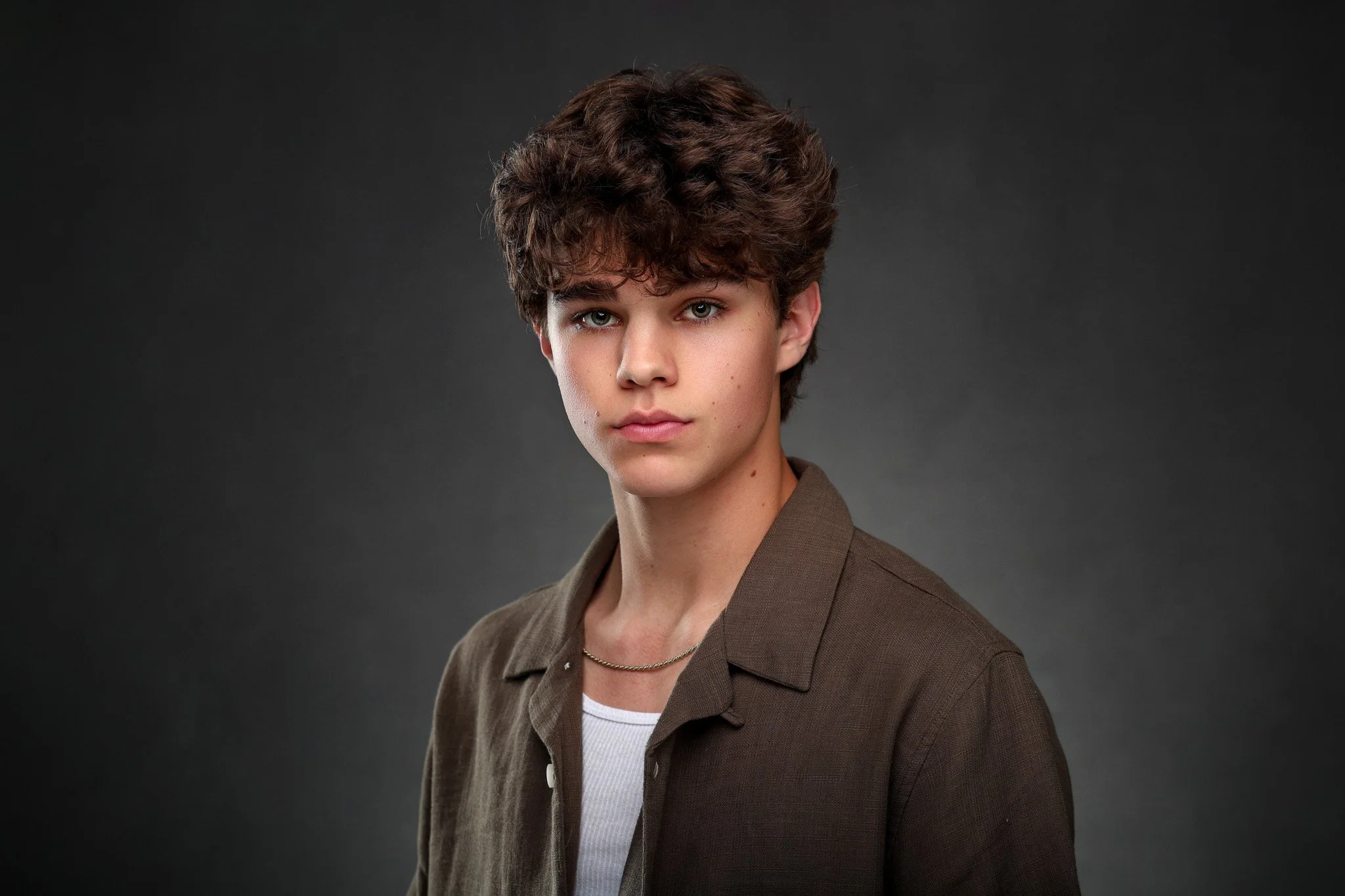 A young woman with short, wavy brown hair, wearing a brown jacket over a white shirt and a thin gold chain, posed against a dark gray background.