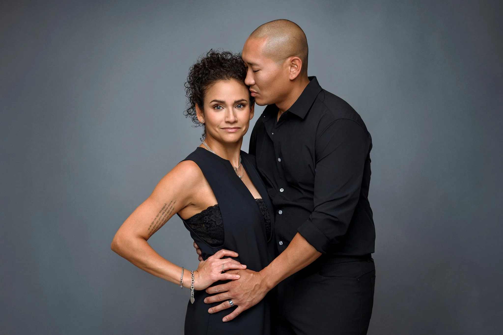 A man and woman in black clothing are posed closely together against a gray background, with the man gently holding the woman's waist and the woman looking at the camera with a neutral expression.