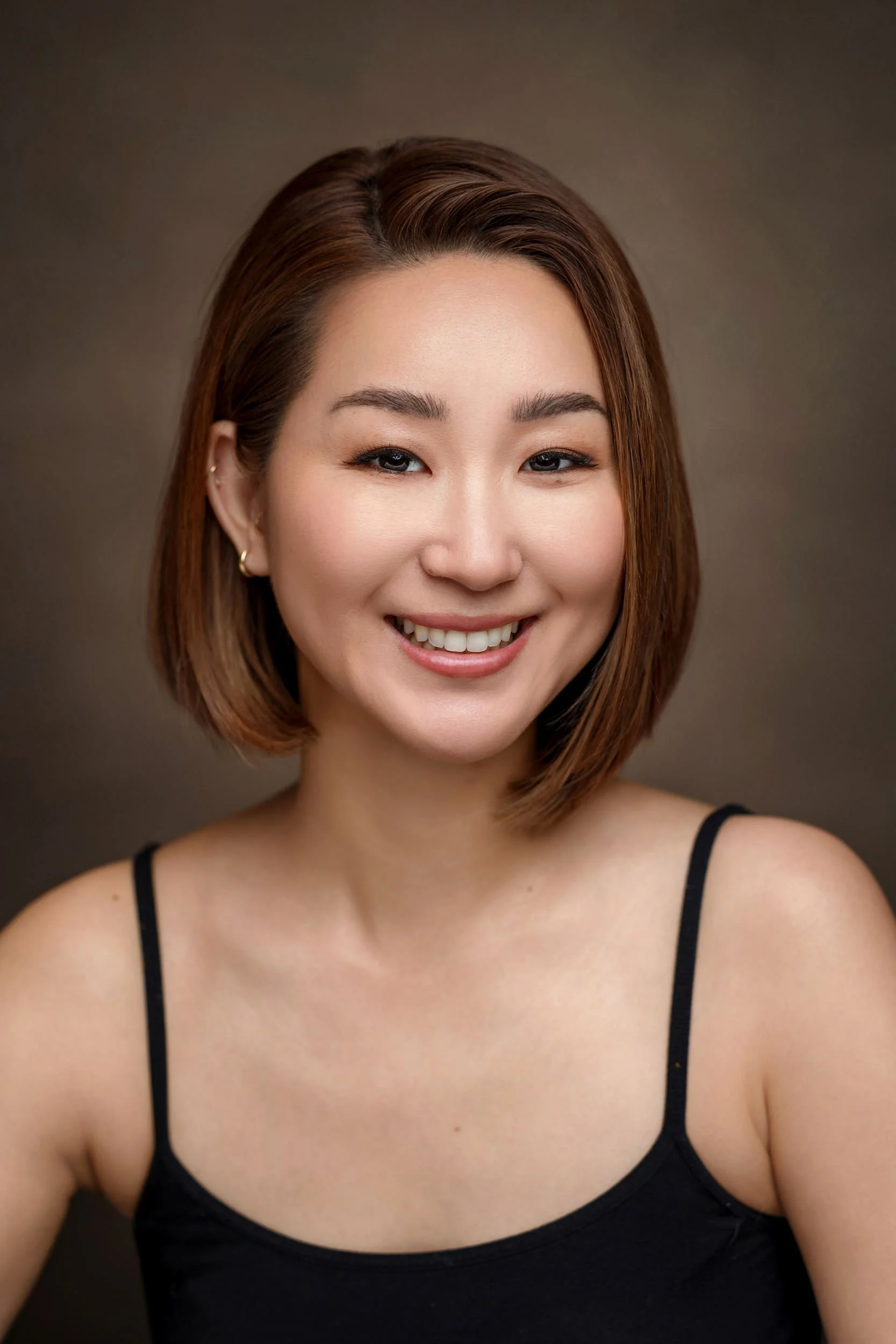 Close-up portrait of a young woman with short brown hair, smiling, wearing a black spaghetti strap top, set against a plain dark background.