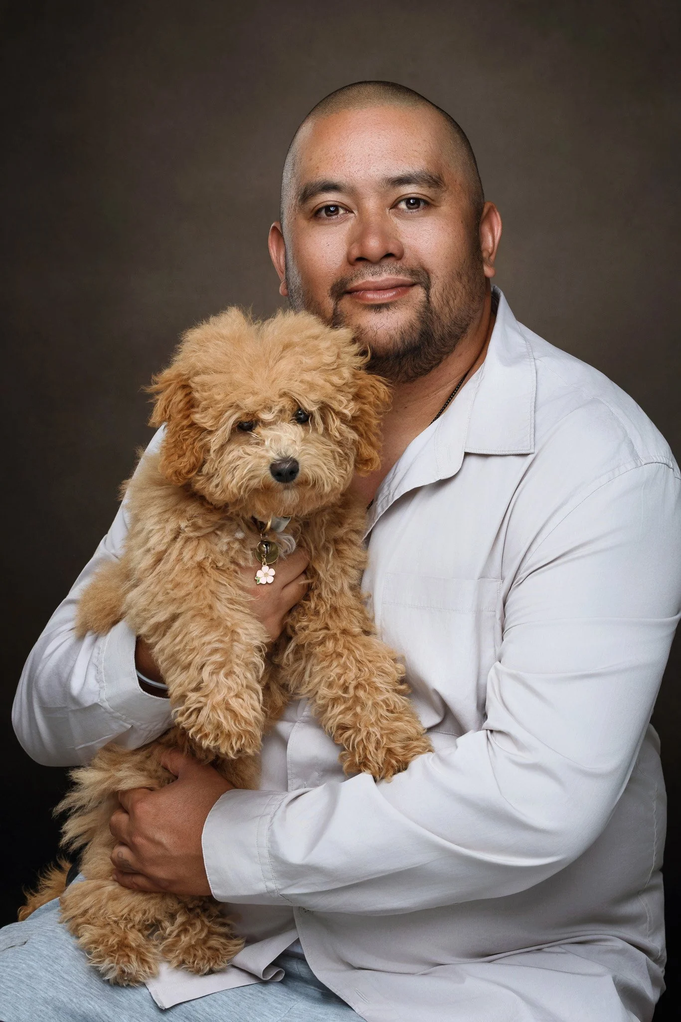 A man with a shaved head and beard holding a fluffy, light brown poodle dog against a dark background.