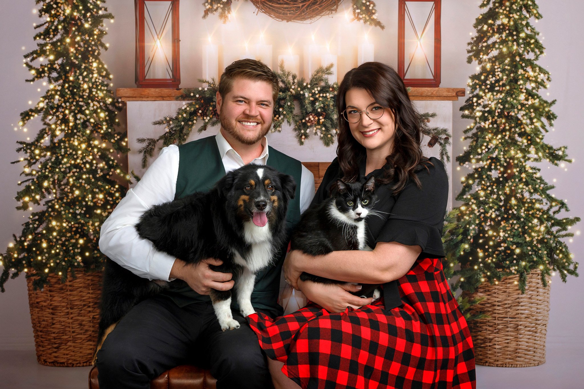 A smiling couple sitting on a bench holding dogs, with a festive Christmas backdrop including decorated trees, wreaths, candles, and lanterns.