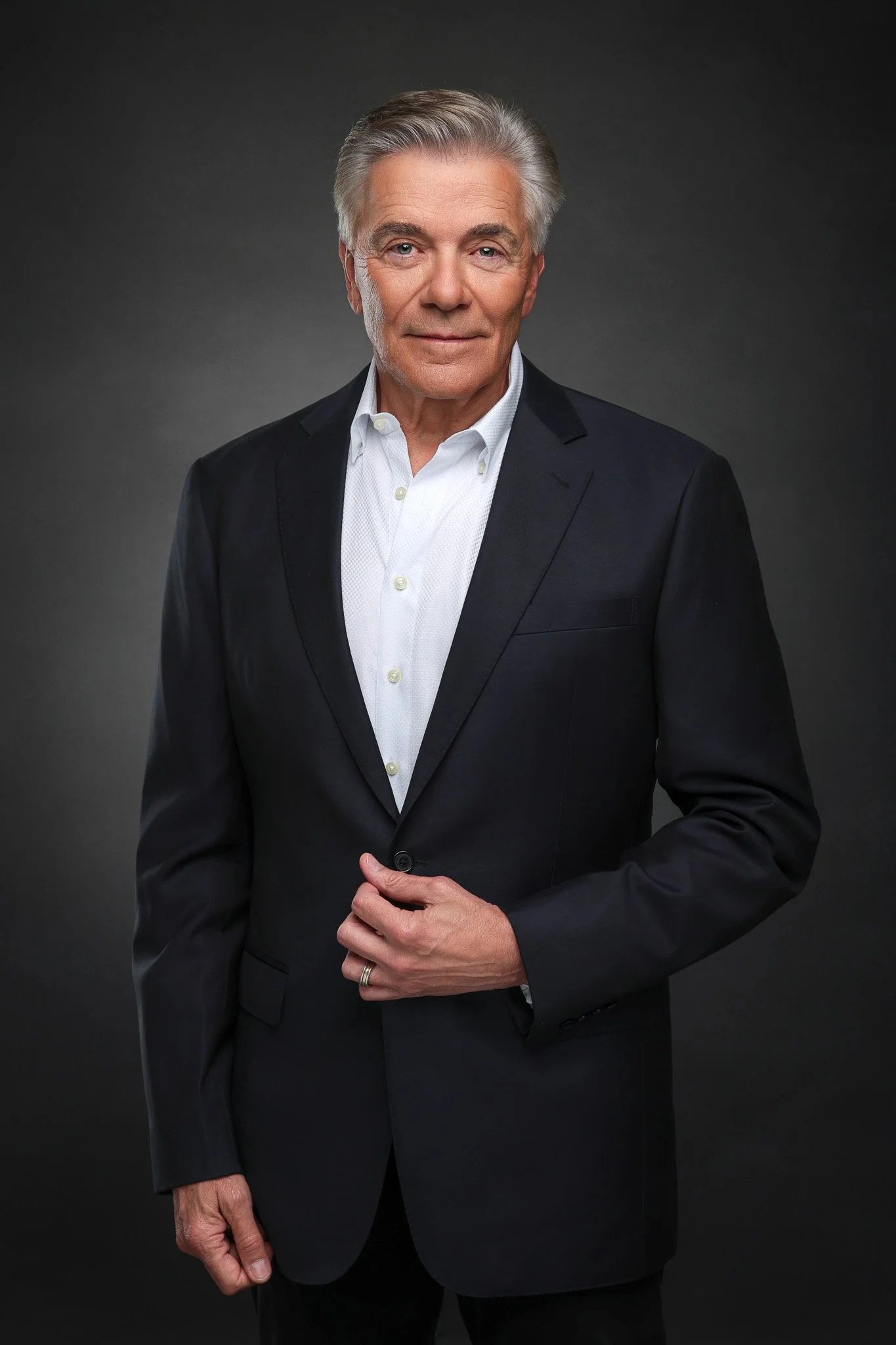 An elderly man with gray hair, wearing a black suit and white dress shirt, stands against a dark gray background, looking directly at the camera with a slight smile.
