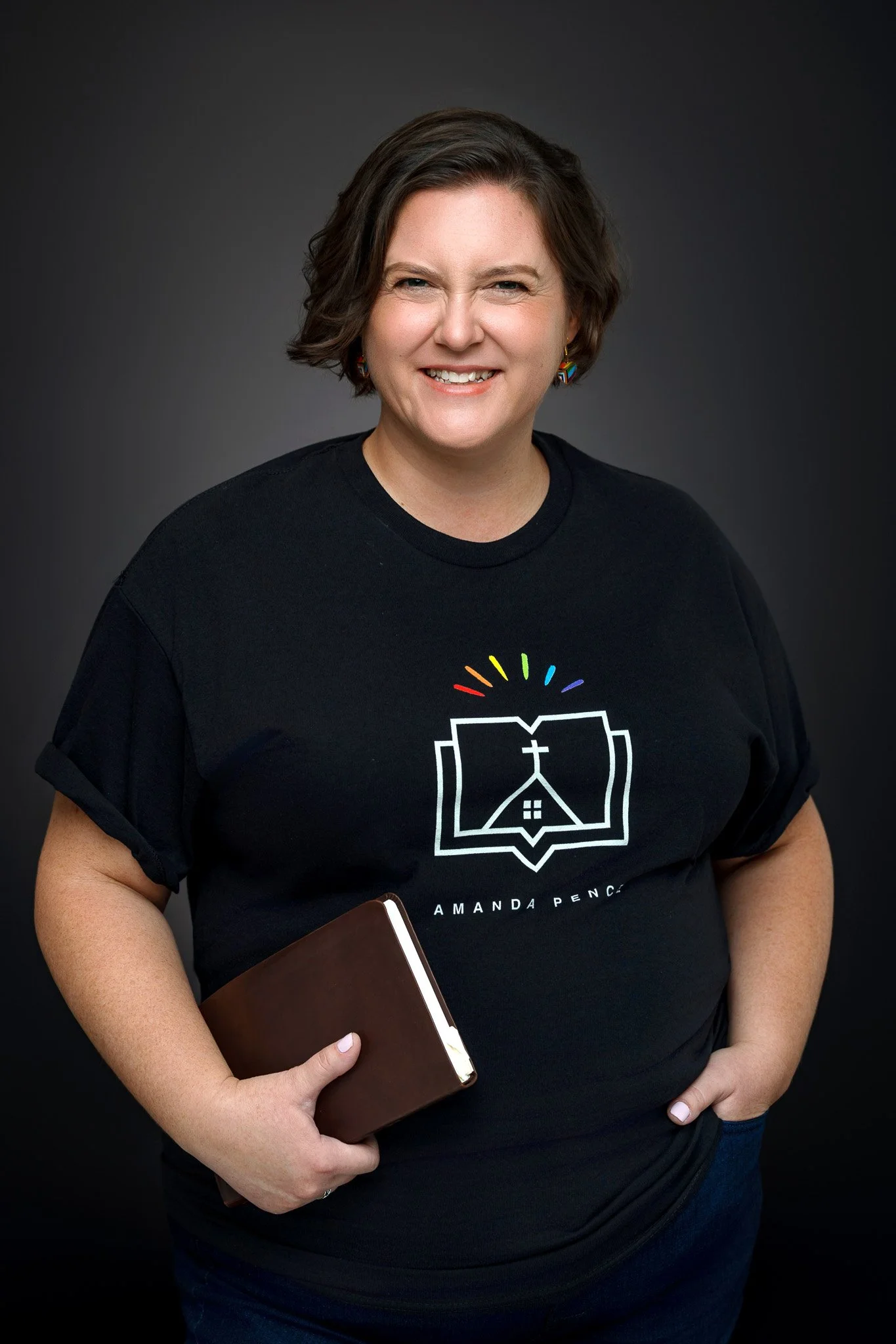 A woman with short brown hair smiling, wearing a black T-shirt with a colorful logo, holding a closed brown notebook, against a dark background.