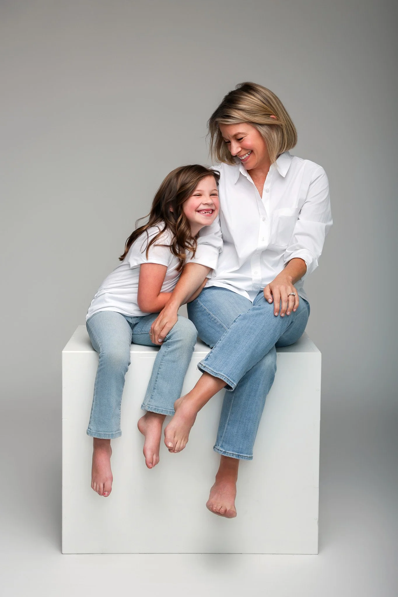 A woman and a young girl sitting on a white cube against a plain gray background, happily laughing and hugging, wearing white shirts and light blue jeans, with their legs dangling over the edge of the cube.