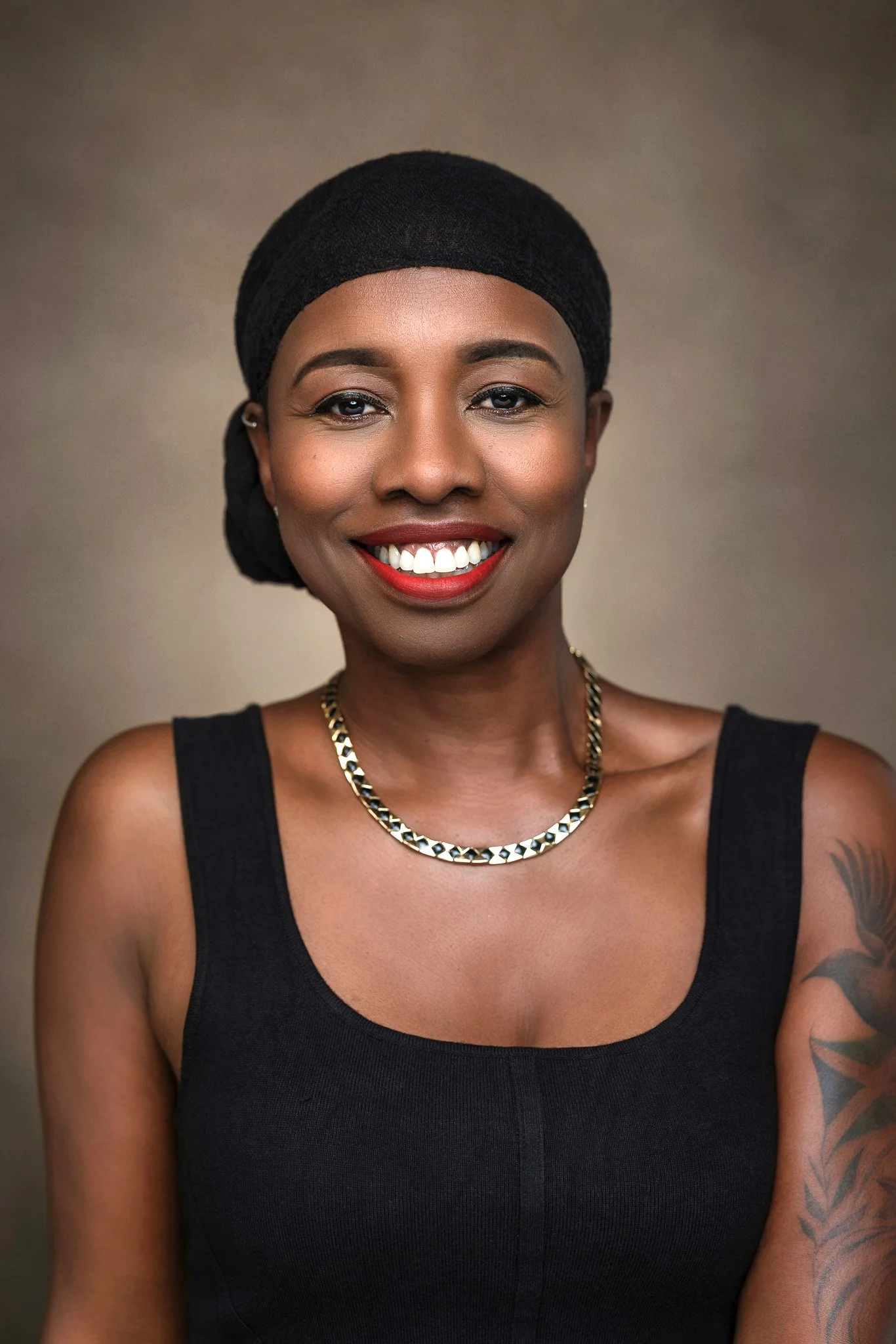 A smiling Black woman with short hair wearing a black headwrap, a black sleeveless top, a gold necklace, and a tattoo on her upper left arm, against a plain background.
