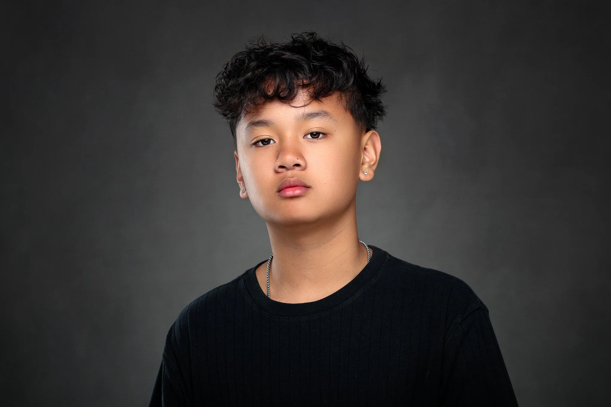 Portrait of a person with short, curly dark hair, wearing earrings, a silver necklace, and a black shirt against a gray background.