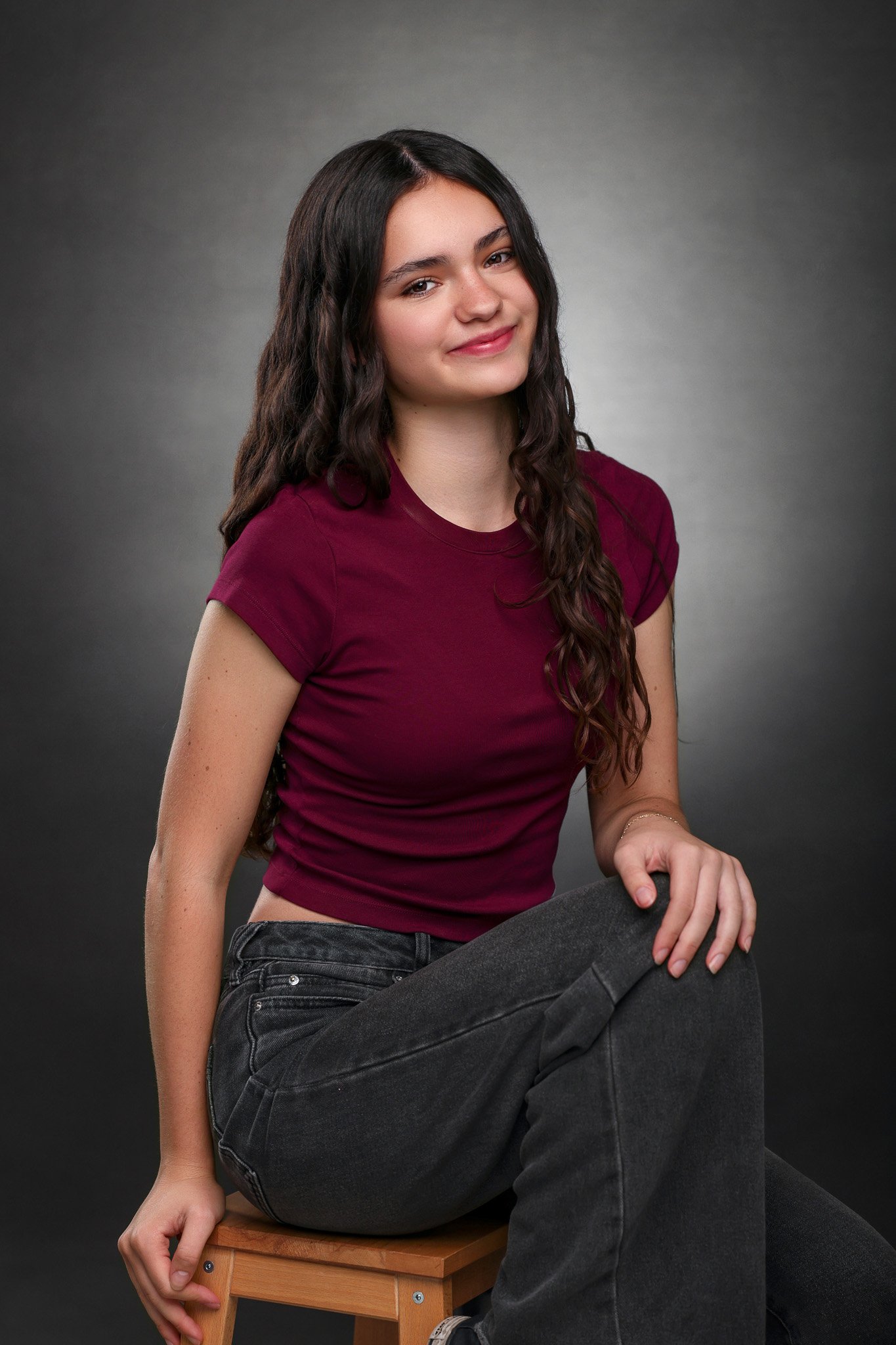 Young woman with long dark curly hair, wearing a burgundy T-shirt and black jeans, sitting on a wooden stool against a gray background.