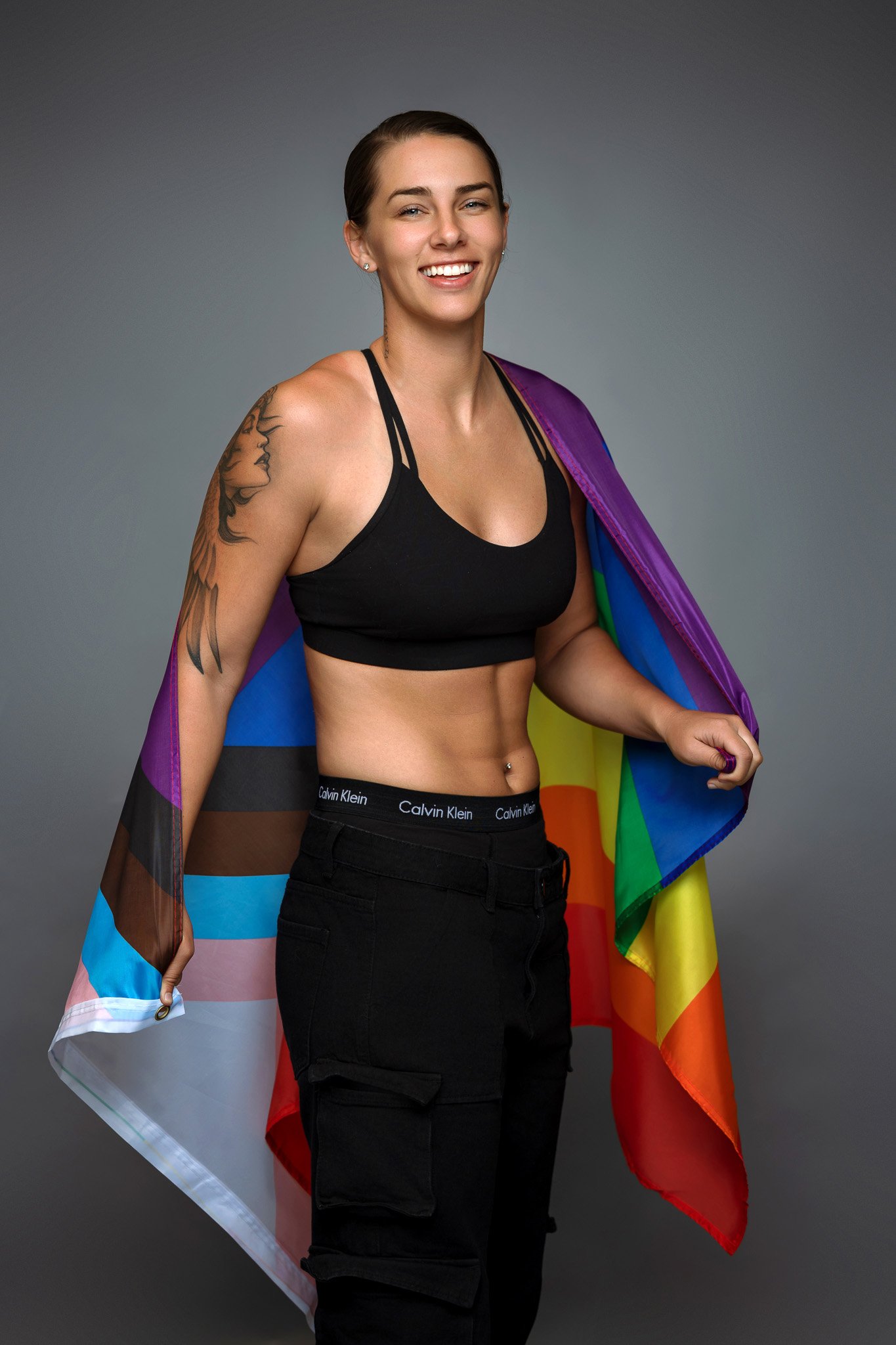 A smiling woman with a rainbow pride flag draped over her shoulders, wearing black athletic wear and cargo pants, standing against a gray background.