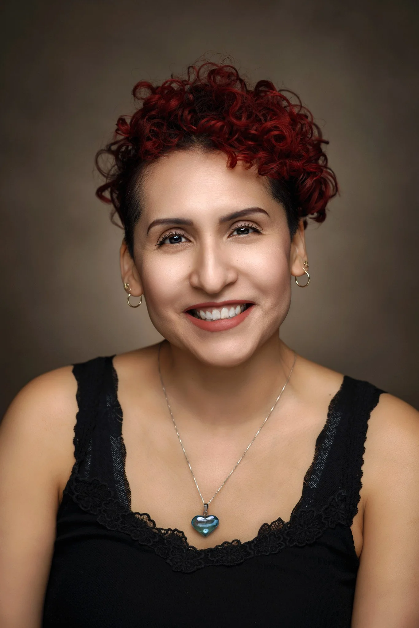 Close-up portrait of a woman with short, curly red hair, light skin, wearing earrings, a silver necklace with a heart-shaped pendant, and a black lace top, smiling against a neutral background.