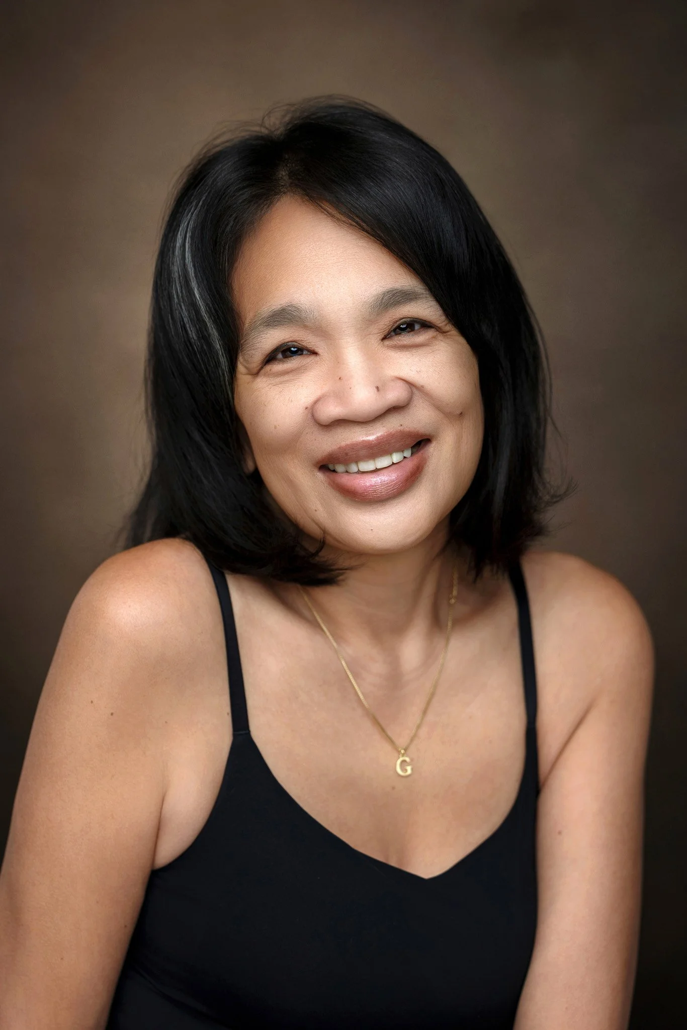 Close-up portrait of a smiling woman with black hair, wearing a black spaghetti strap top and a gold necklace with a 'G' pendant, against a brown background.