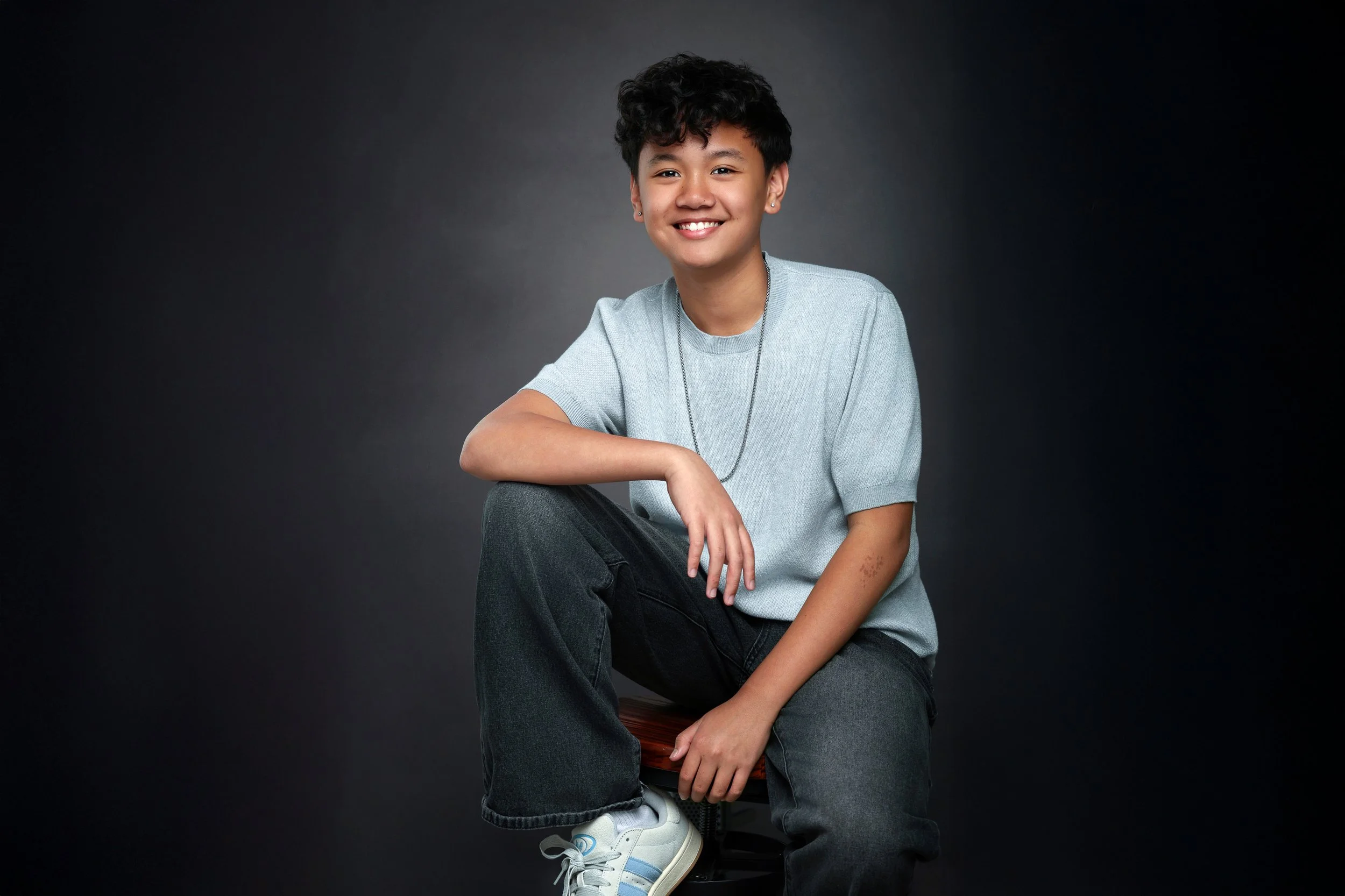 Young person with curly dark hair smiling, dressed in a light gray T-shirt and dark jeans, sitting on a stool against a dark gray background.