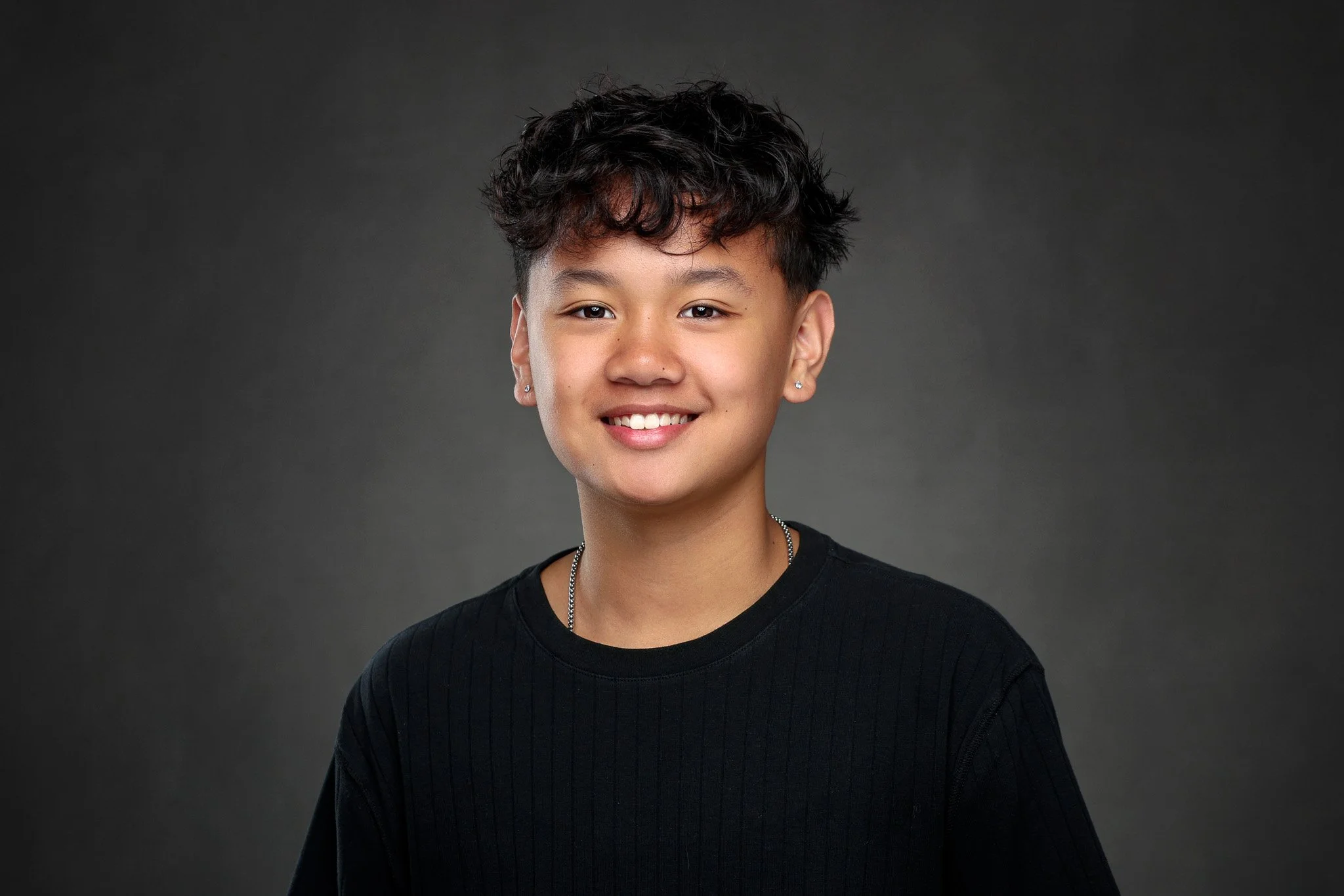 Portrait of a young woman with short curly black hair, wearing earrings, a black shirt, and a silver necklace, smiling against a dark background.