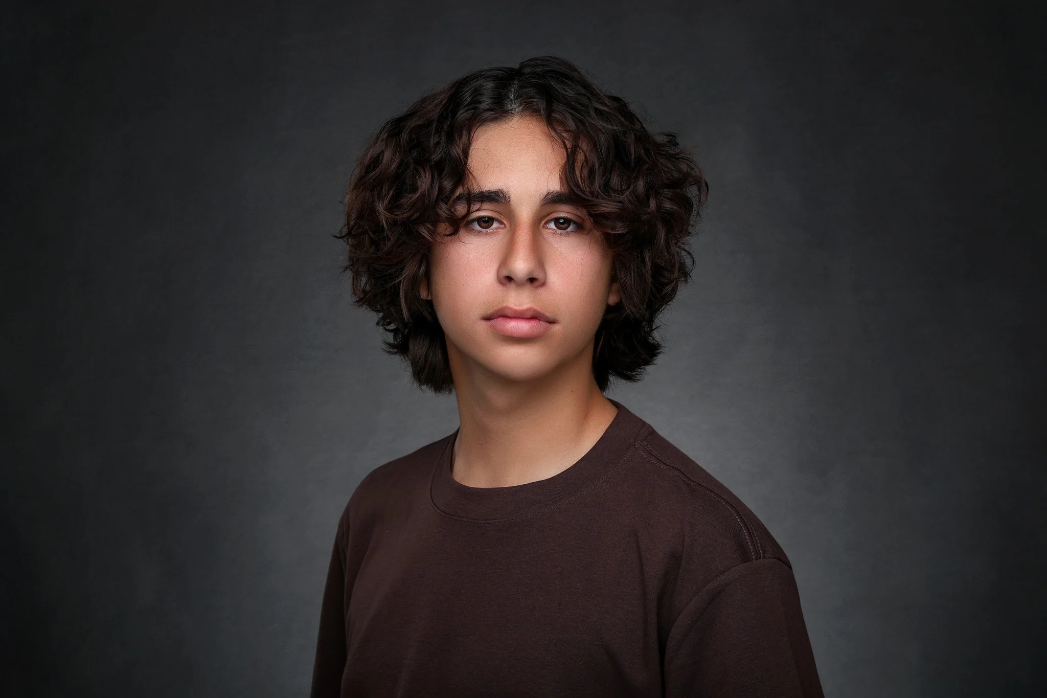 A young person with curly dark hair and light skin looking directly at the camera, wearing a dark sweater, against a dark gray background.