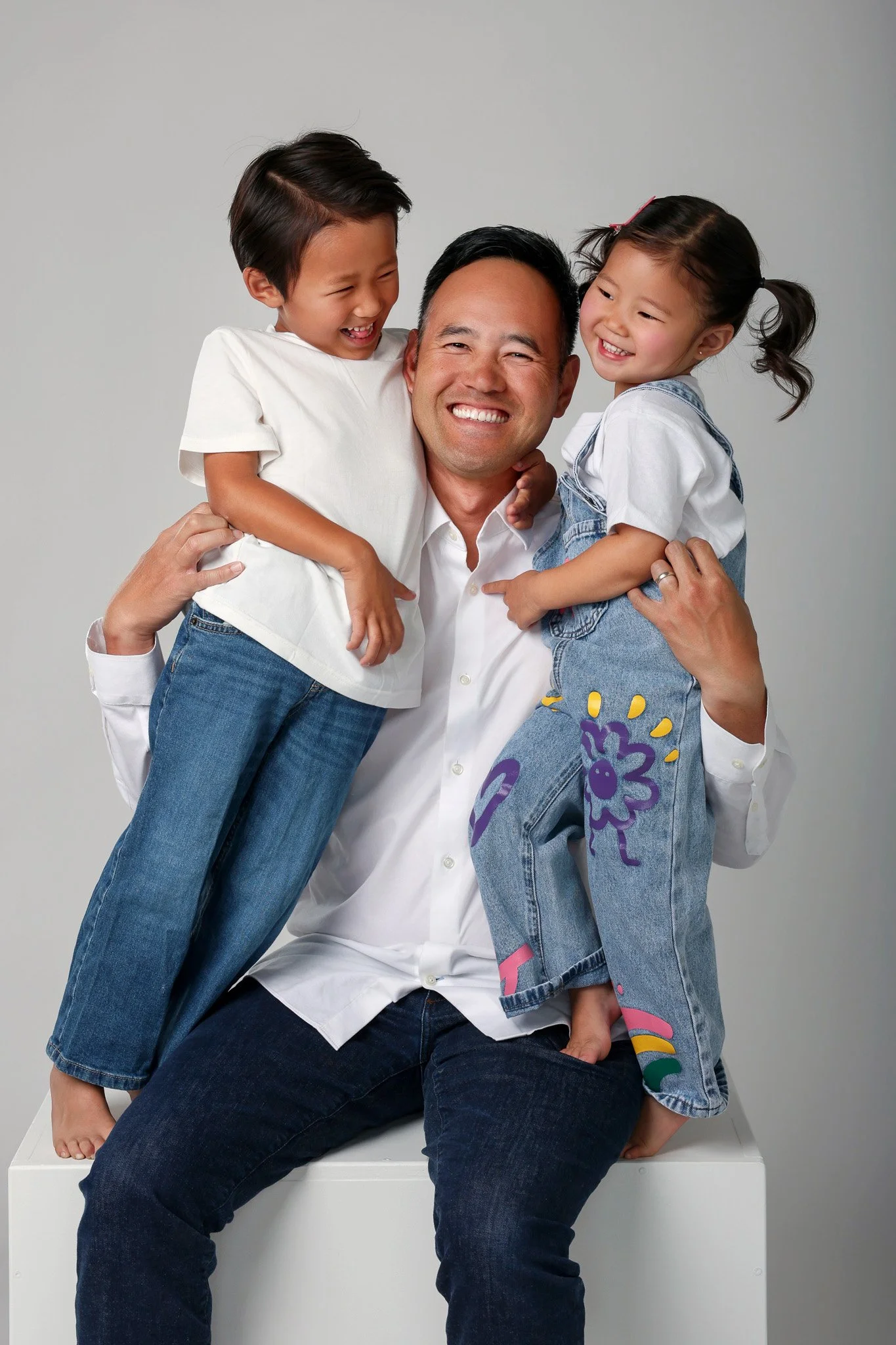 A man sitting on a white box, smiling, with two children on his lap and shoulders, all laughing and hugging each other against a plain gray background.