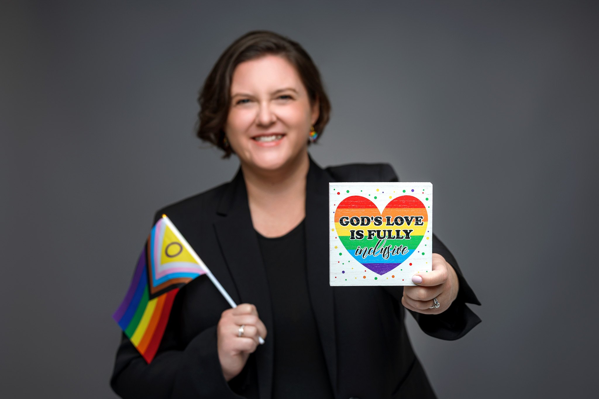 Woman smiling, holding a small rainbow pride flag and a sign with a rainbow heart and the text "God's love is fully inclusive" against a plain gray background.