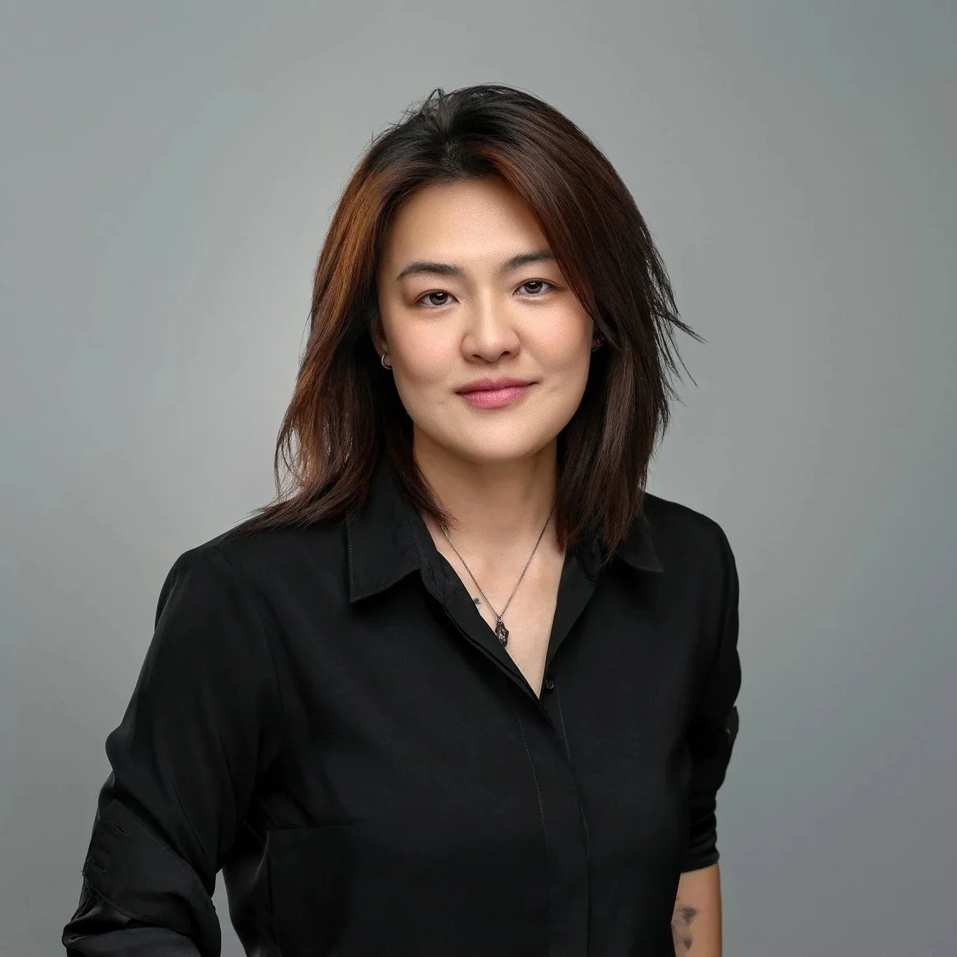Portrait of a young woman with shoulder-length brown hair, wearing a black shirt and silver necklace, standing against a gray background.