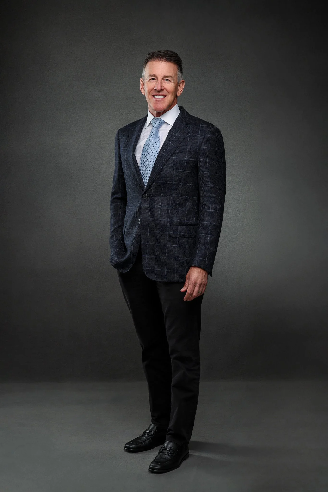 A man wearing a dark checkered blazer, white shirt, light blue polka dot tie, black pants, and black shoes, standing against a dark gray background, smiling and looking at the camera.