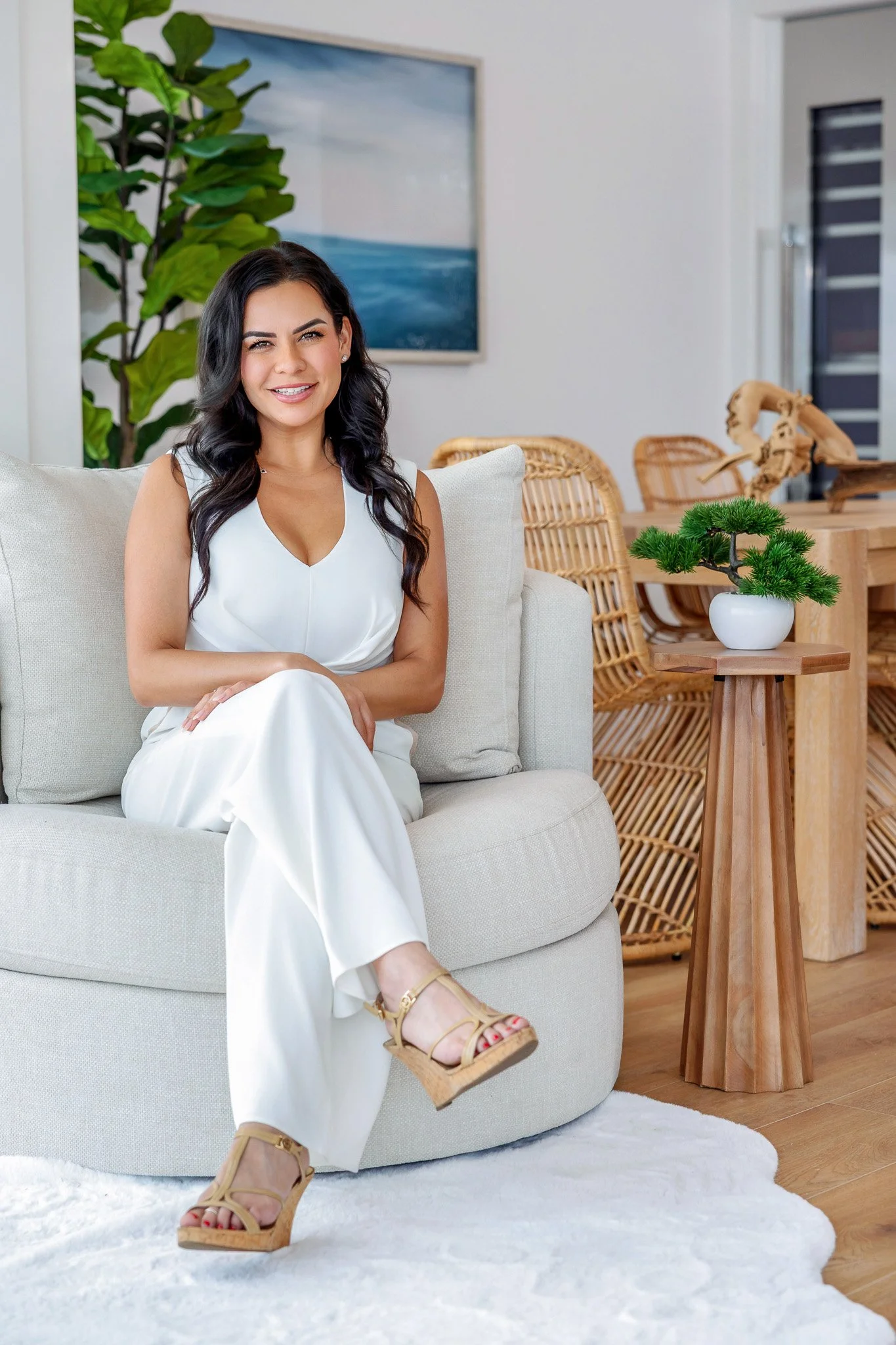 A woman sitting on a beige sofa with a white dress, a potted green plant on a wooden side table, a large framed painting of a blue ocean scene on the wall, and wicker chairs in the background.