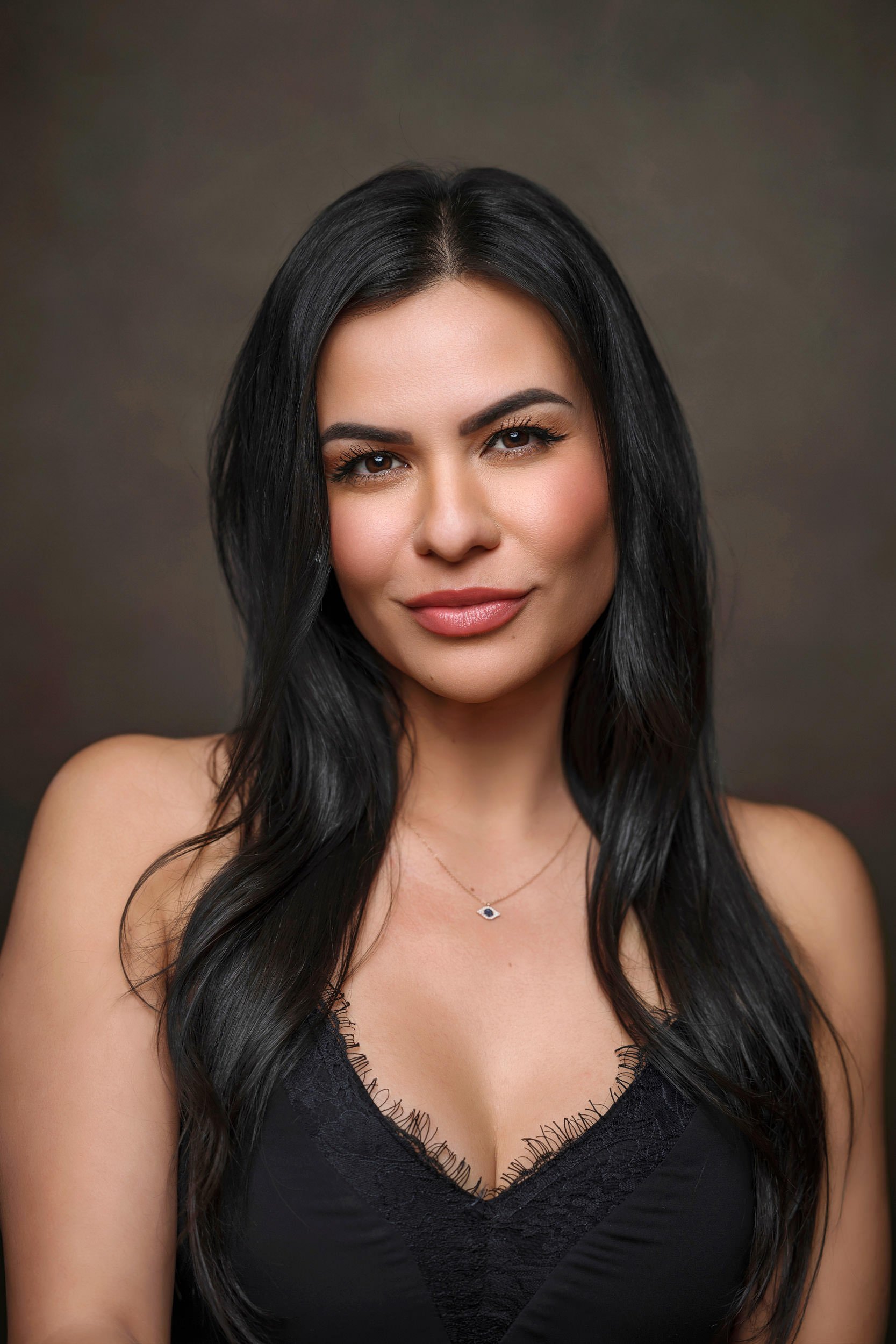Close-up of a woman with long black hair, wearing a black lace top and a delicate eye-shaped necklace, smiling slightly against a neutral background.